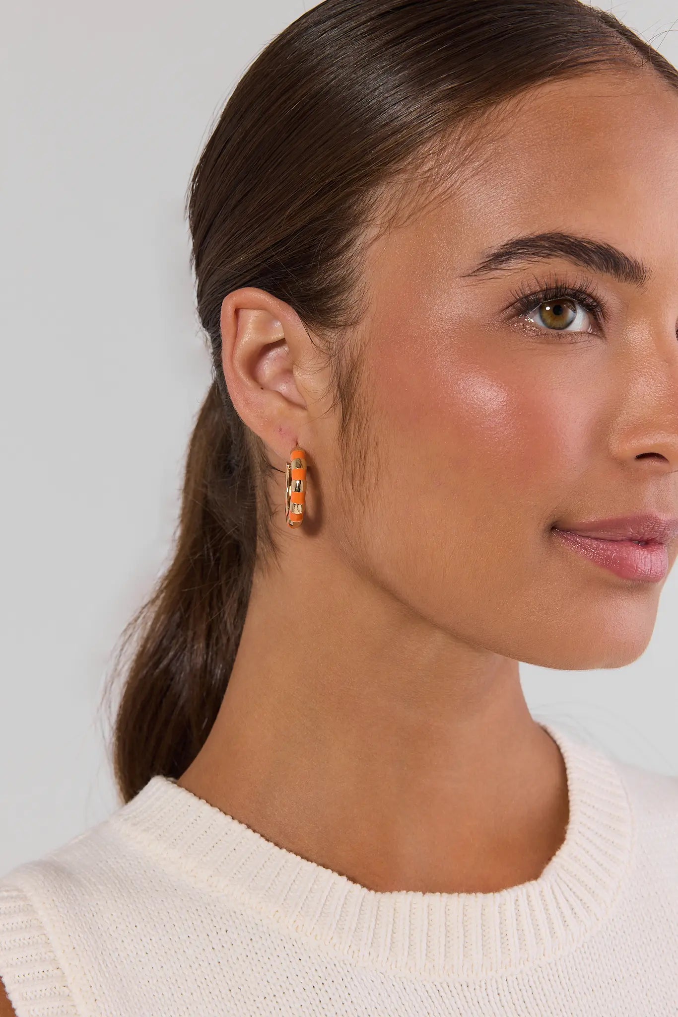 A close-up of a woman with her hair in a ponytail wearing a small gold hoop earring with orange and white geometric accents, paired with a cream ribbed knit top against a light gray background.