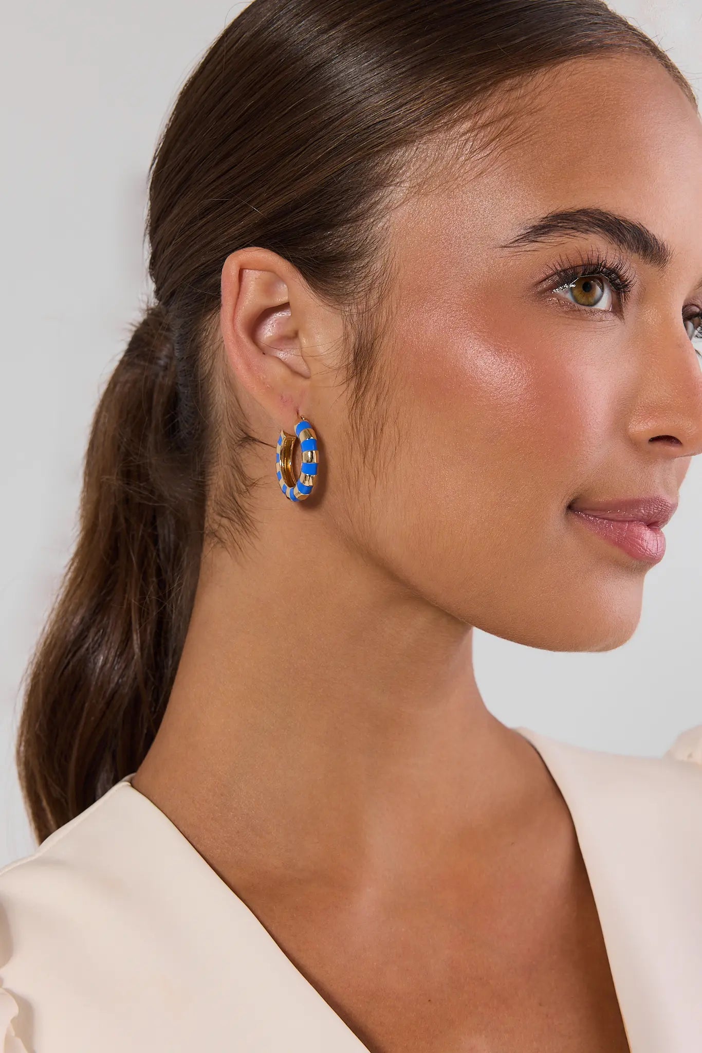 A close-up side profile of a woman with dark brown hair in a ponytail, wearing a vibrant blue and orange patterned hoop earring, against a light gray background.