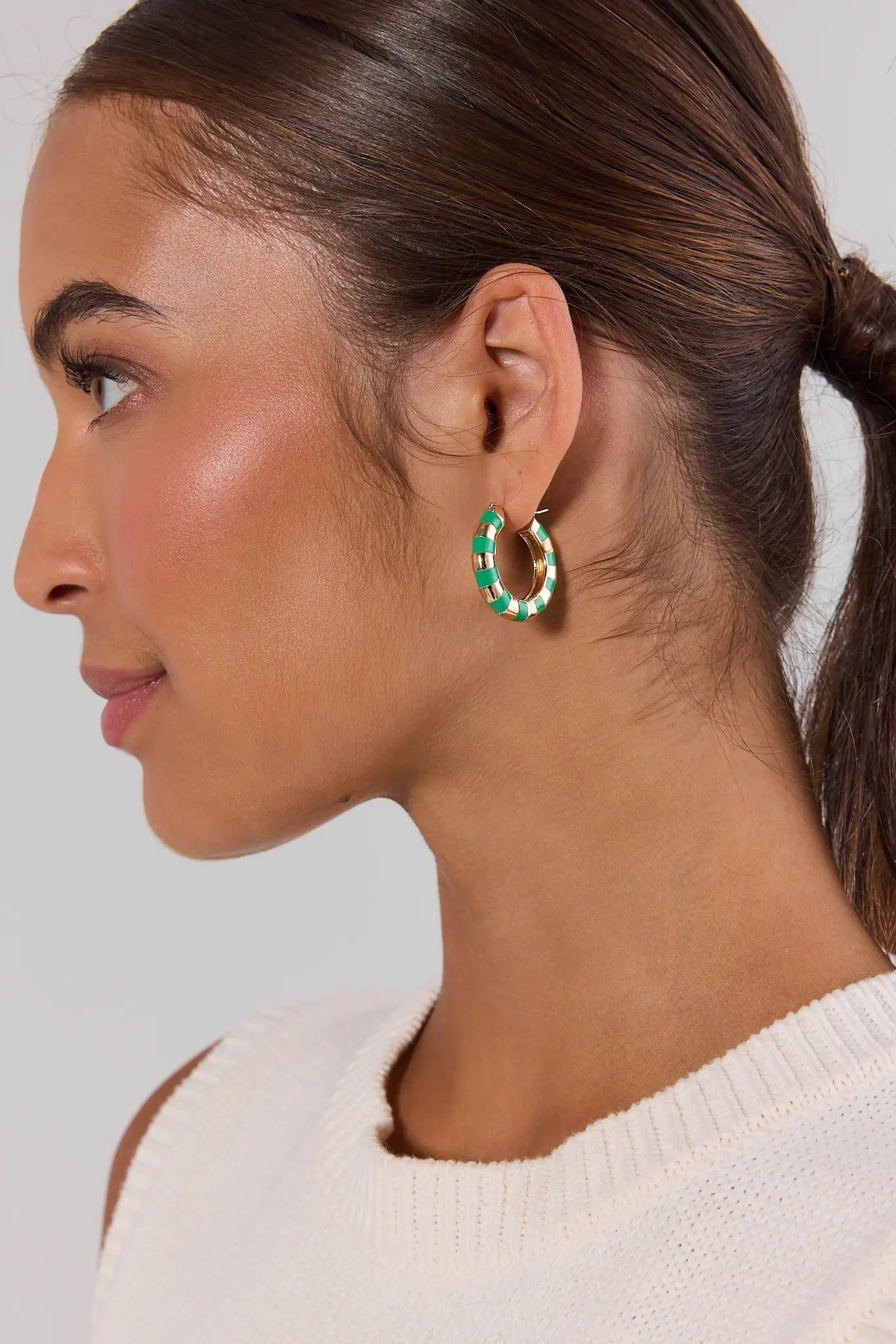 A close-up side profile of a woman wearing a small gold hoop earring with green and white marbled accents, paired with a white knit top against a light gray background.