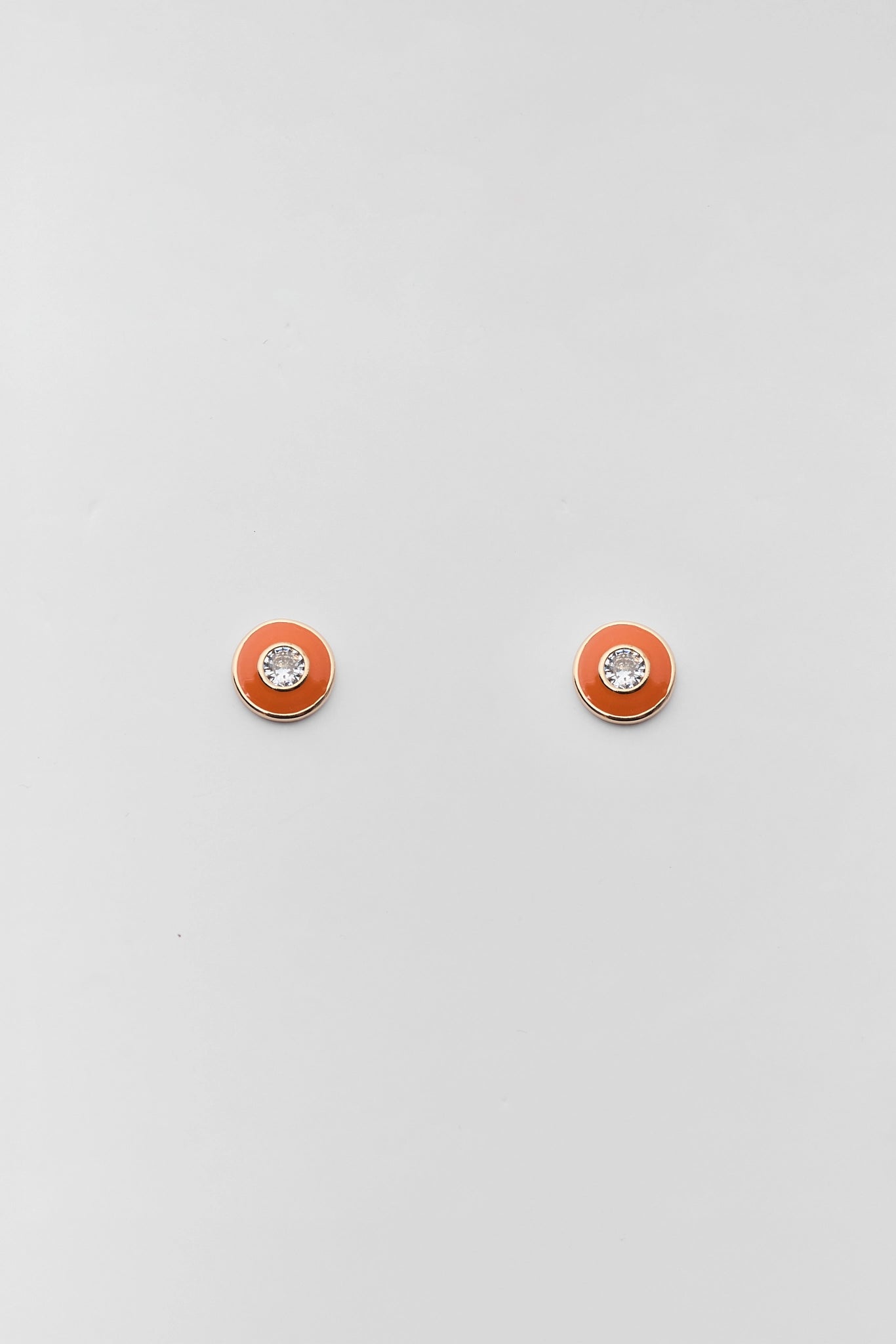A pair of small round stud earrings with a vibrant orange-red enamel face, a silver-tone bezel, and a clear crystal center, displayed on a light gray background.