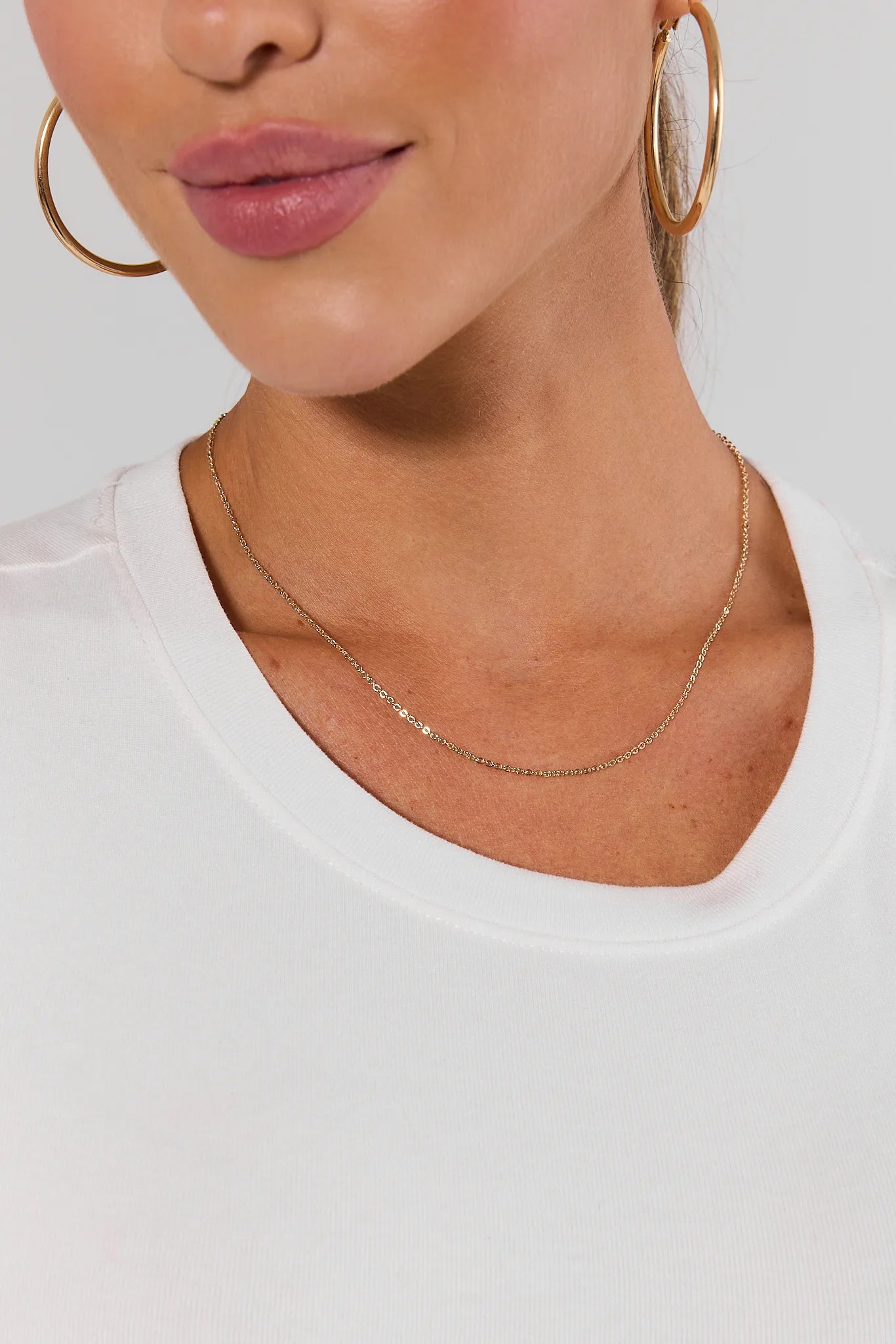 A close-up of a model wearing a delicate gold chain necklace, gold hoop earrings, and a white crew-neck top against a light gray background.