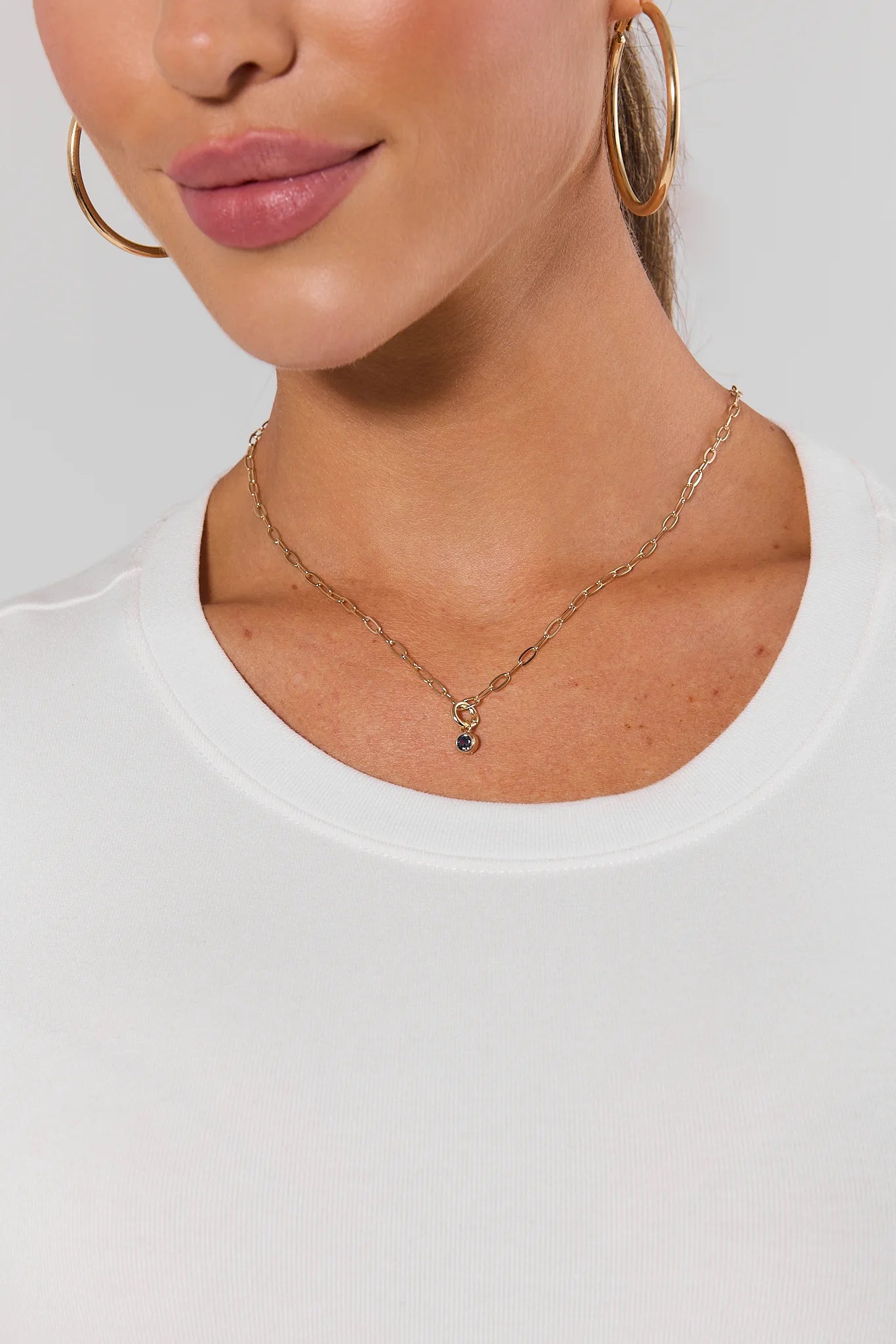 A close-up of a model wearing a delicate gold chain necklace with a small round purple gemstone pendant, paired with a white crew-neck top.
