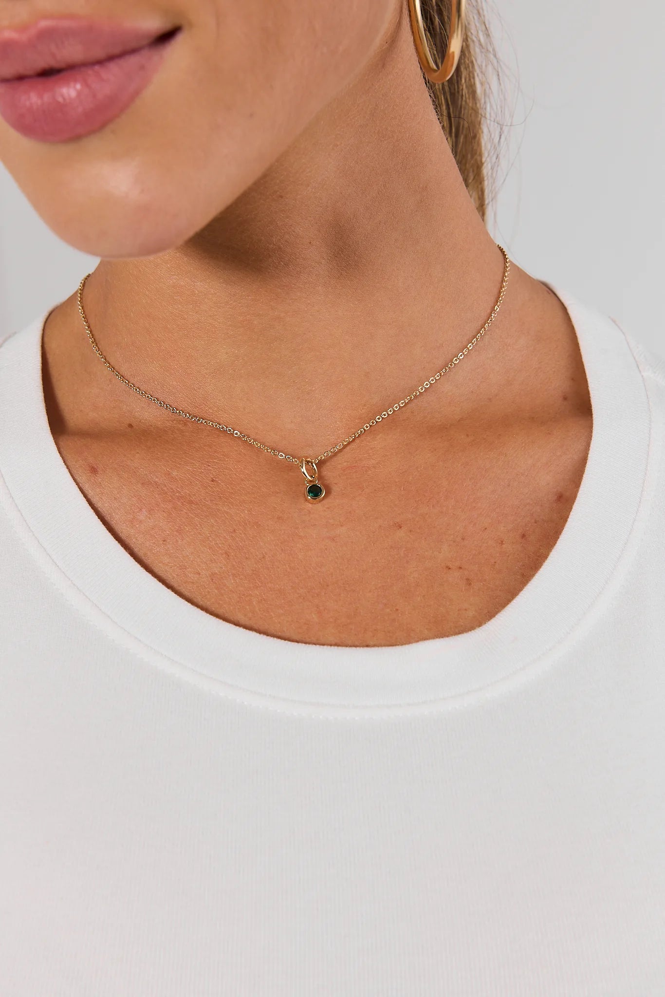 A close-up of a model wearing a delicate gold chain necklace with a small round dark green gemstone pendant, paired with a white crew-neck top.