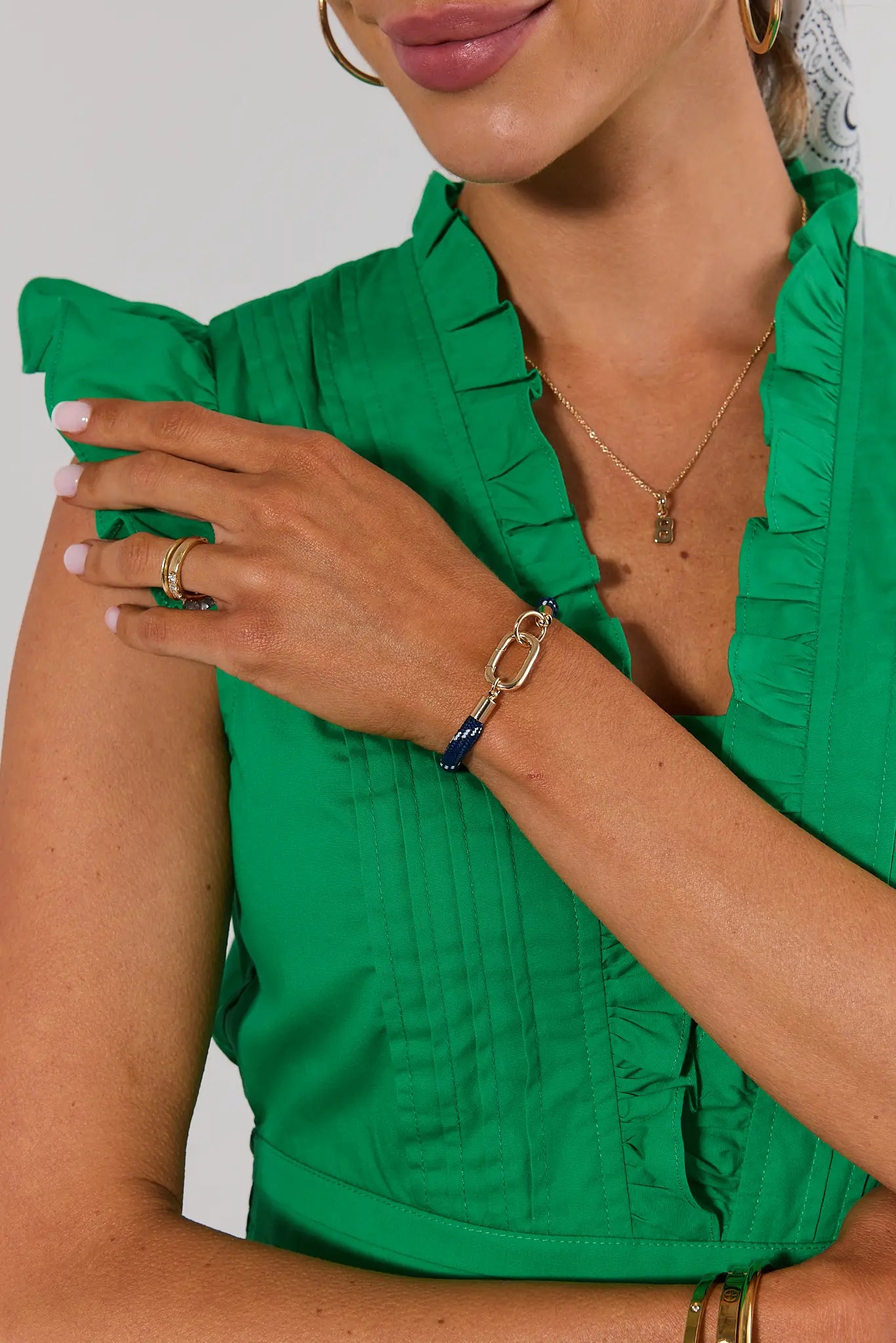 A close-up of a model wearing a bright green ruffled blouse, accessorized with gold rings, a gold chain necklace, and a blue and gold link bracelet.