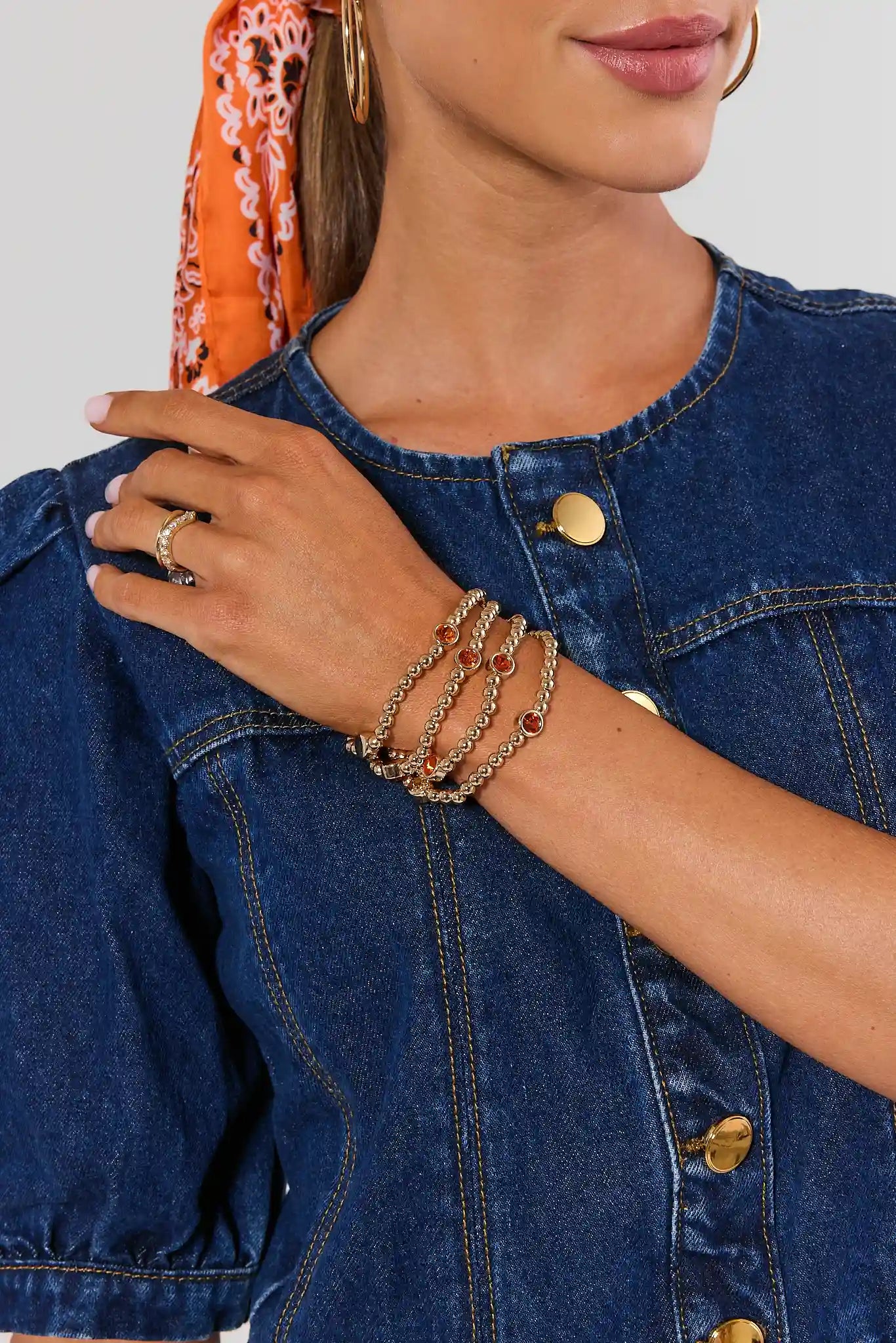 A close-up of a model wearing a blue denim jacket with gold-tone buttons, layered gold chain bracelets on one wrist, and a patterned orange scarf.