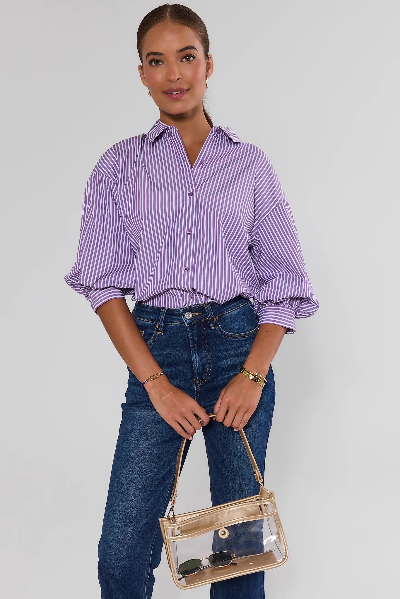 A woman modeling a lavender and white vertically striped button-up shirt with voluminous puff sleeves, paired with dark blue jeans and holding a clear tote bag with a beige frame.