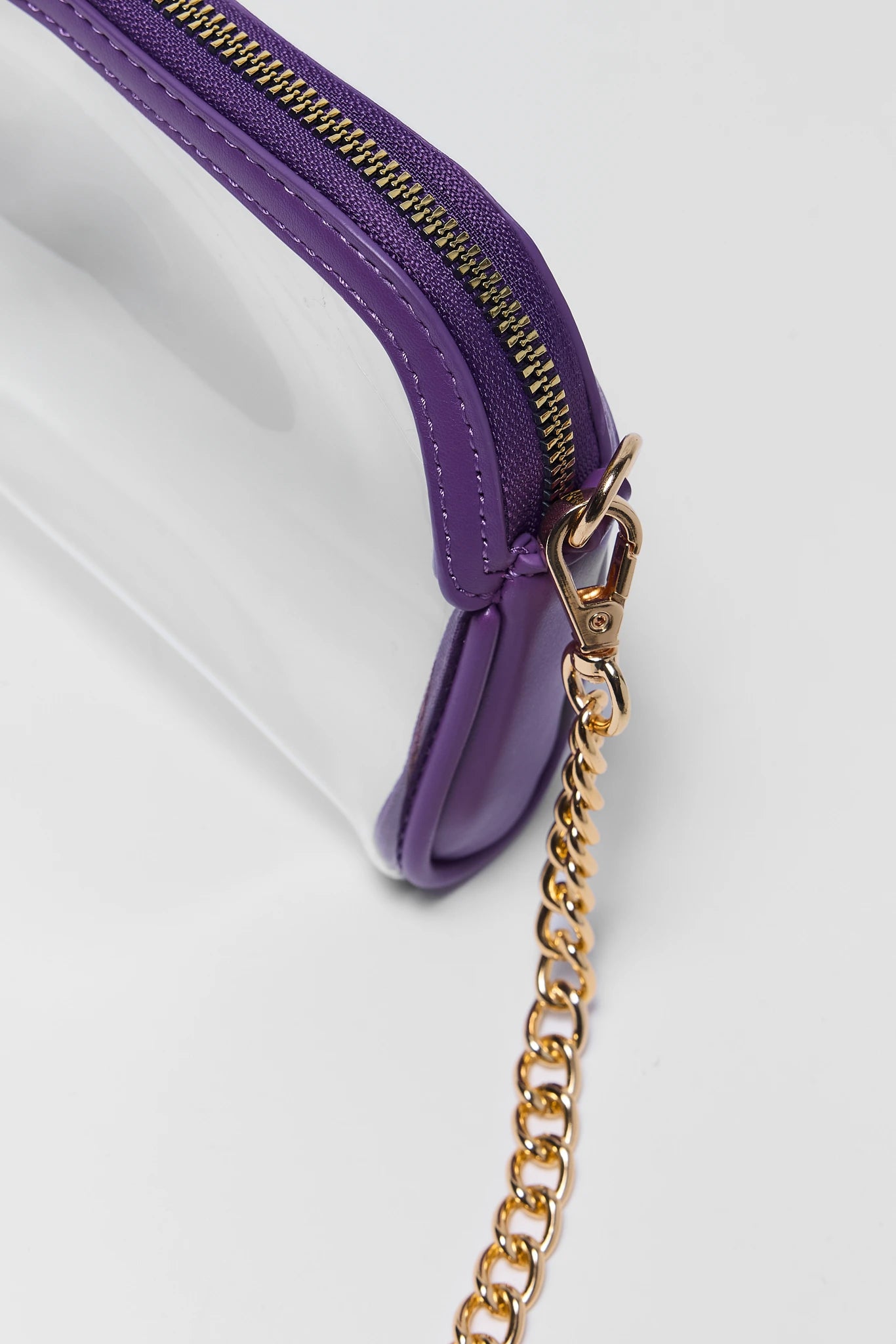 Close-up of a clear plastic pouch with a purple zipper and a gold-tone chain strap on a light gray background.