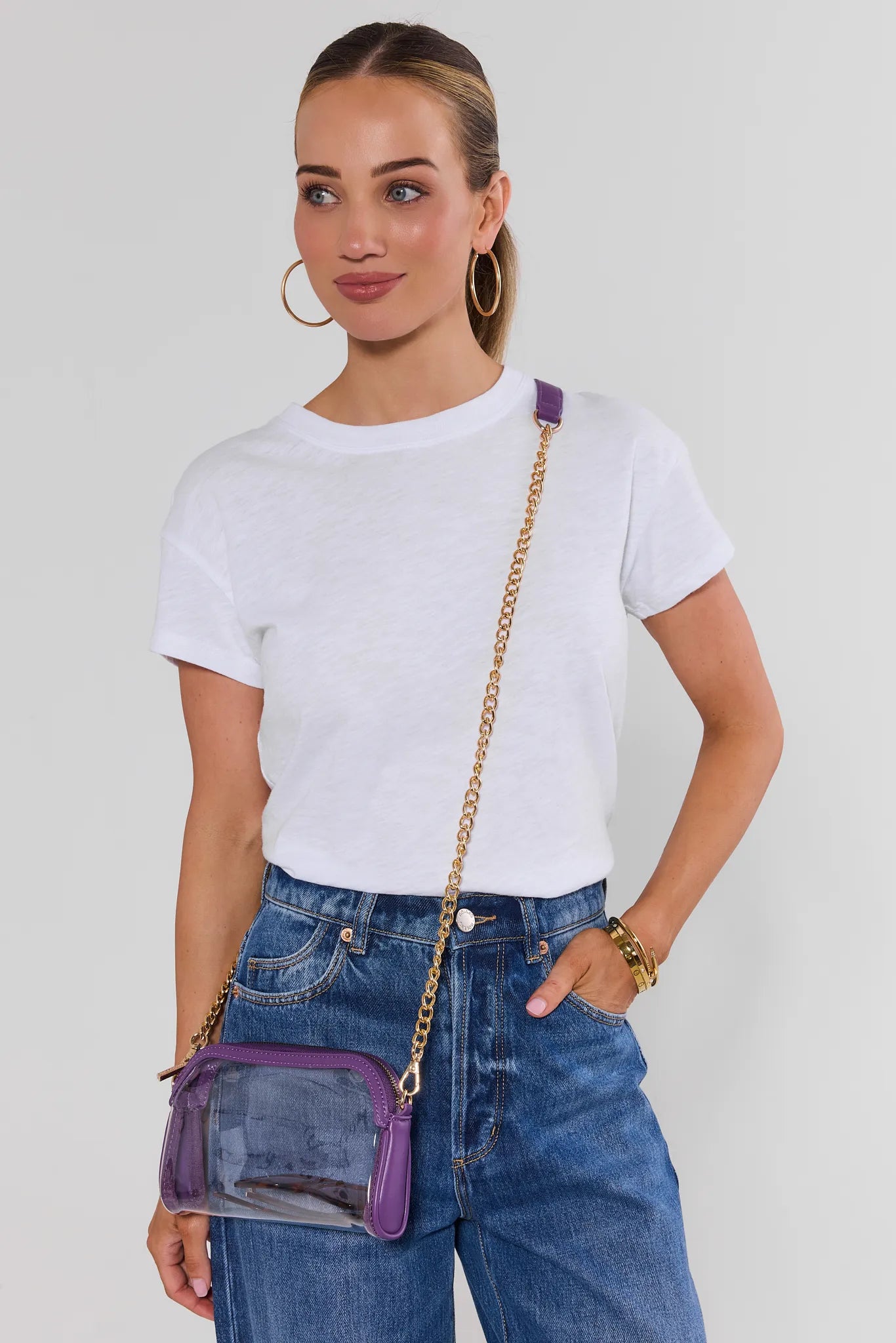 A woman wearing a white t-shirt and blue jeans models a purple and clear plastic crossbody bag with a gold chain strap against a light gray background.