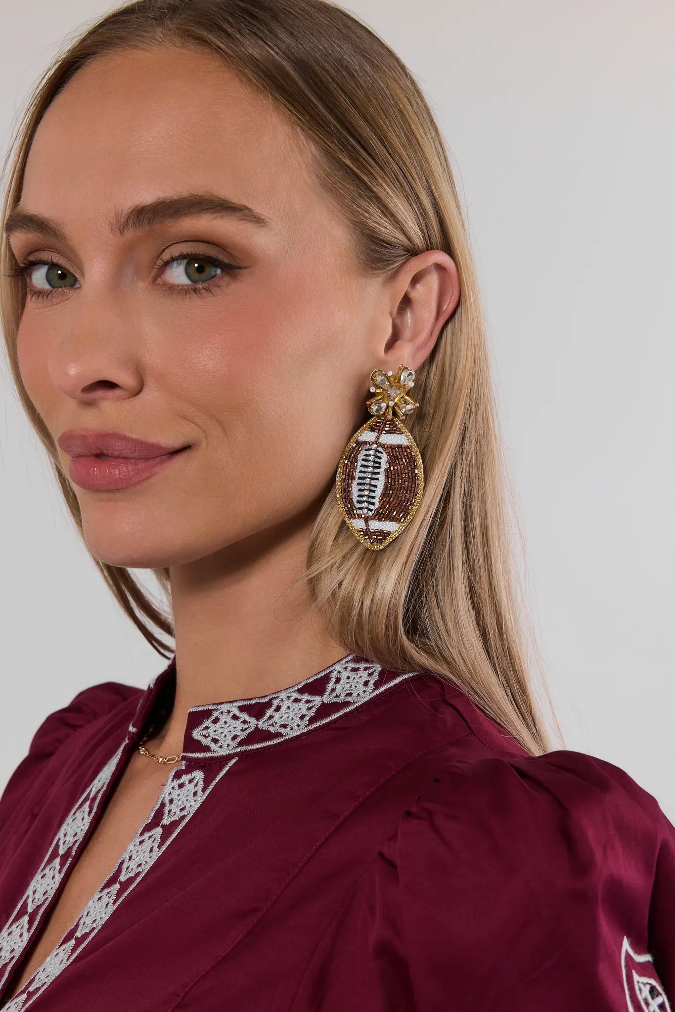 A close-up of a woman wearing a maroon top with white embroidered trim and a large football-shaped dangling earring with gold and white detailing.
