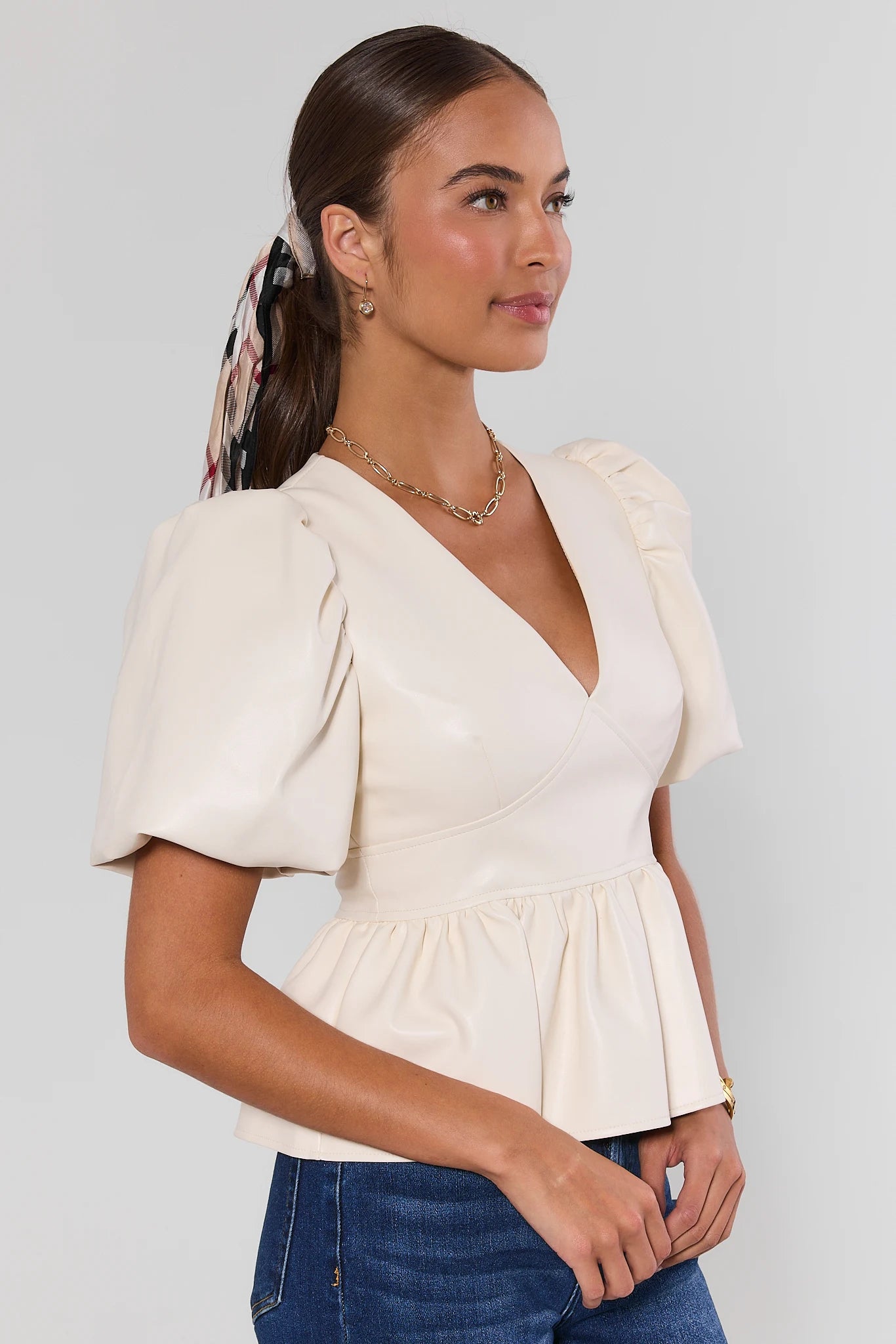 A woman wearing a cream-colored peplum top with puff sleeves and a deep V-neckline, paired with blue jeans, photographed against a neutral light gray background.