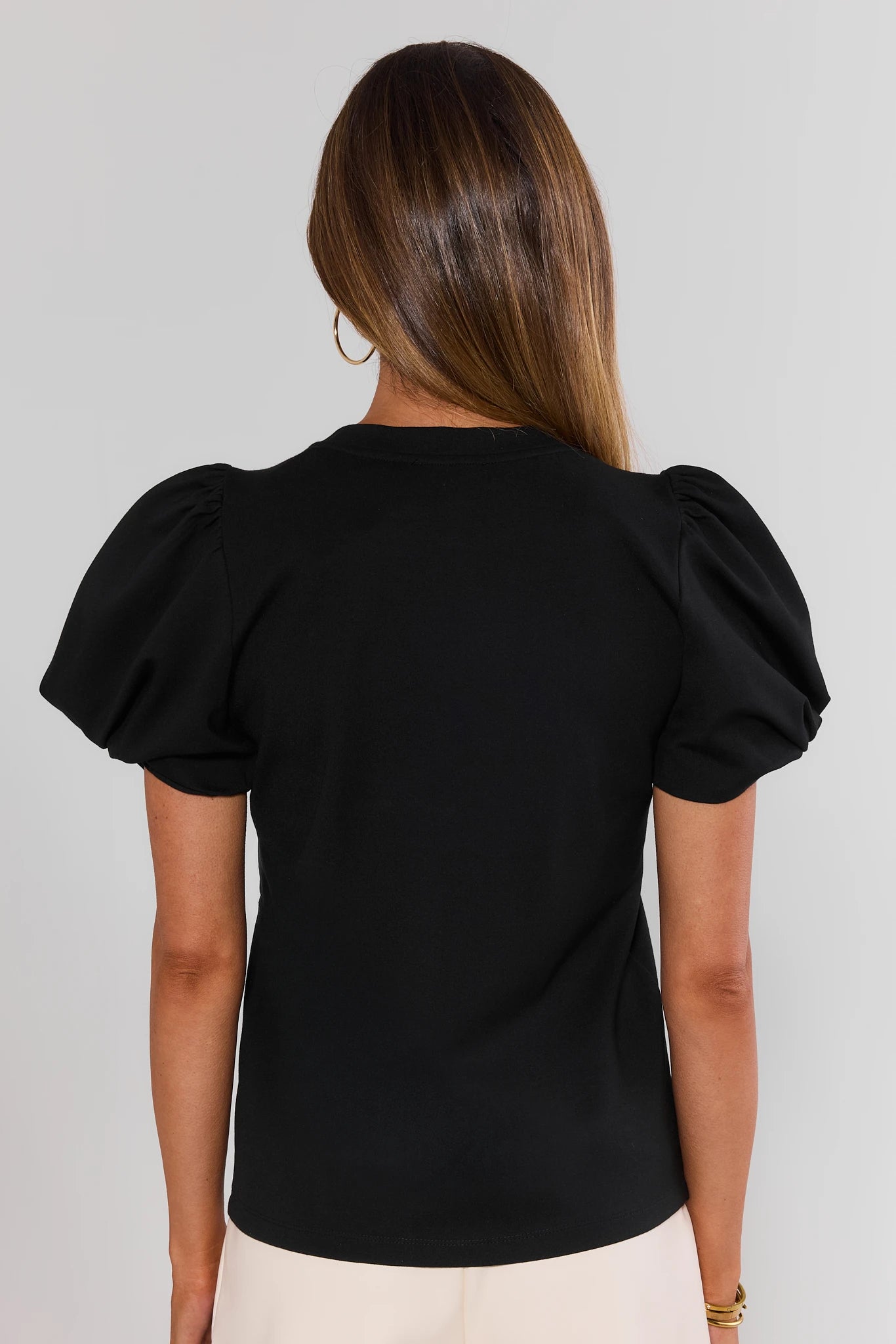 A woman shown from behind wearing a black short-sleeve top with voluminous puff sleeves and white high-waisted trousers, photographed against a gray studio background.