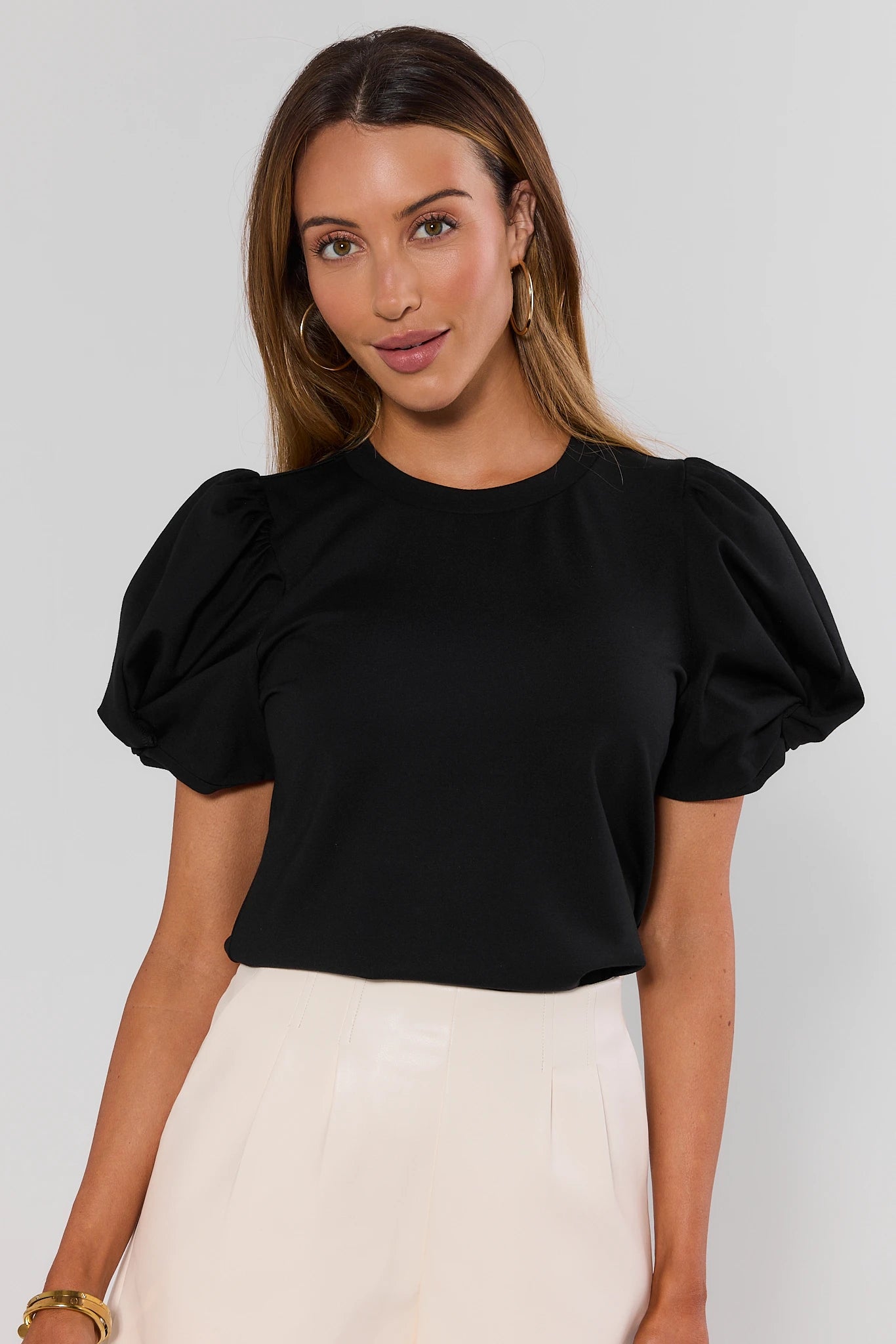 A woman wearing a black short-sleeve top with voluminous puff sleeves and white high-waisted trousers, photographed against a light gray studio background.