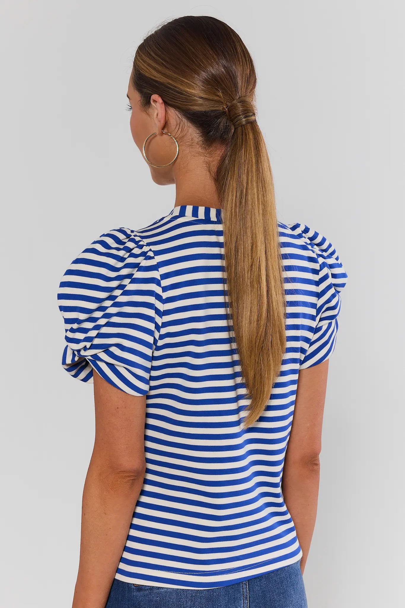 A woman shown from behind wearing a blue and white horizontally striped short-sleeve top with puffed sleeves, shown from the chest up against a light gray studio background.