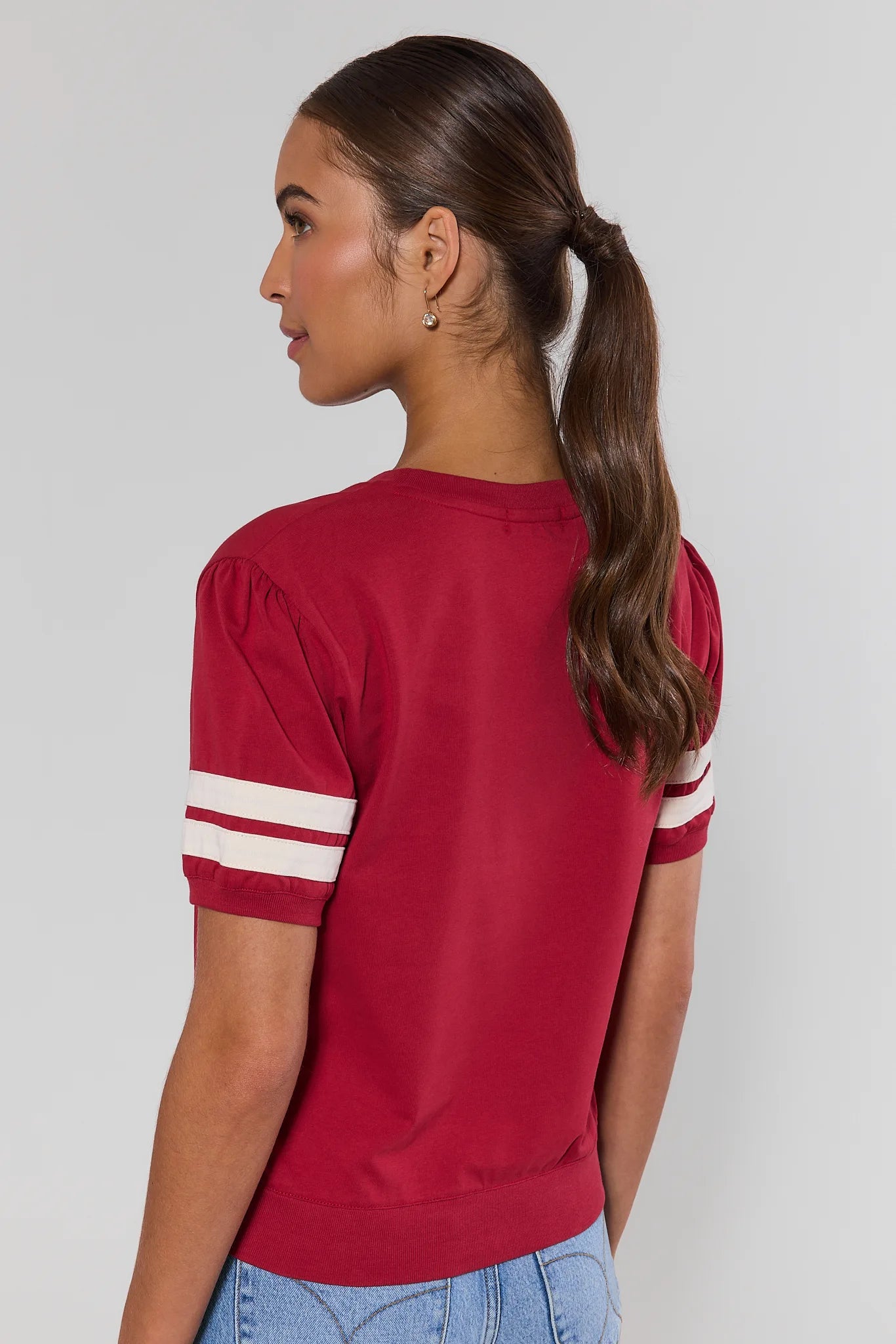 A woman shown from the back wearing a short-sleeve crewneck top in deep red with two white stripes on each sleeve, paired with light blue jeans against a light gray studio background.