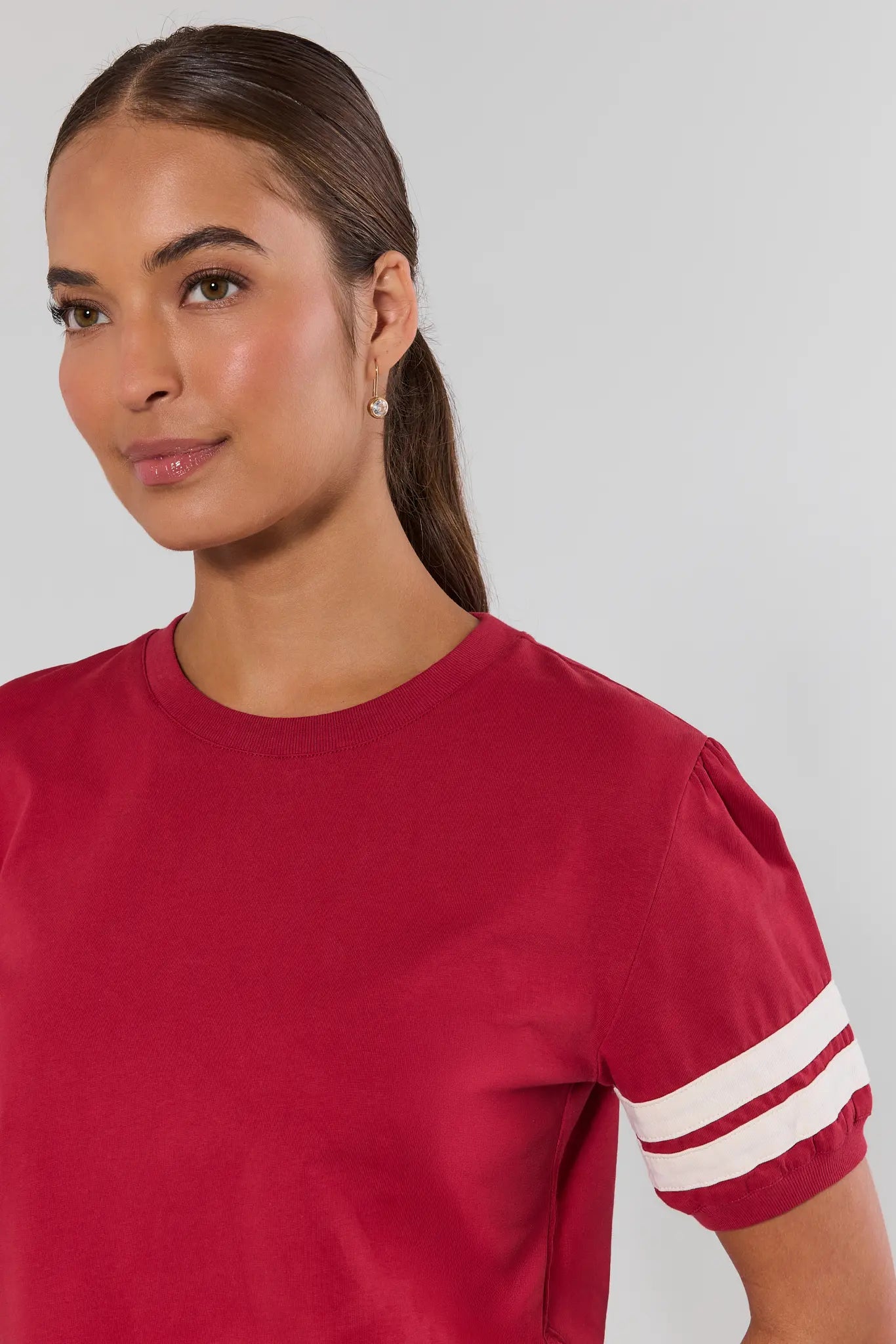 Close up a woman wearing a short-sleeve crewneck top in deep red with white stripes on the sleeves, paired with light blue jeans, against a plain light gray background.