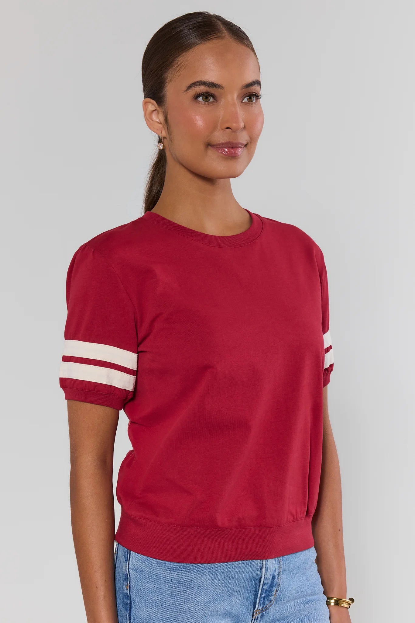 A woman from the side wearing a red short-sleeve crewneck sweatshirt with white stripes on the sleeves, paired with light blue jeans and beige sneakers, against a light gray studio background.