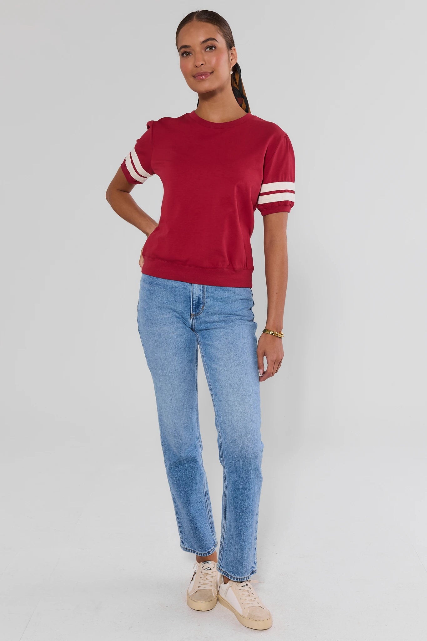A woman modeling a red short-sleeve crewneck sweatshirt with white stripes on the sleeves, paired with light blue jeans and beige sneakers, against a light gray studio background.