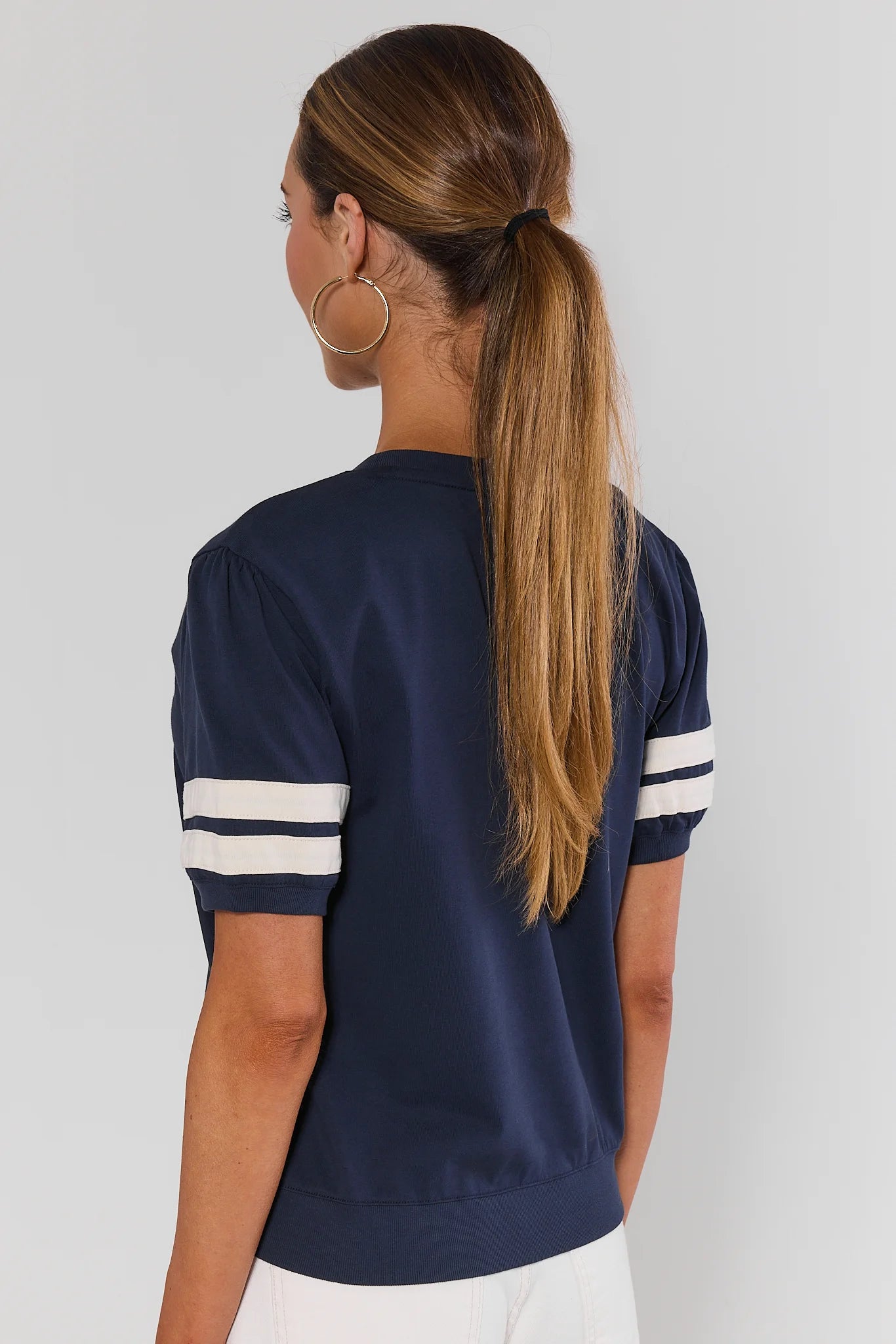 A woman shown from behind modeling a navy blue short-sleeve t-shirt with white stripes on the sleeves, paired with white jeans and white sneakers, against a light gray studio background.