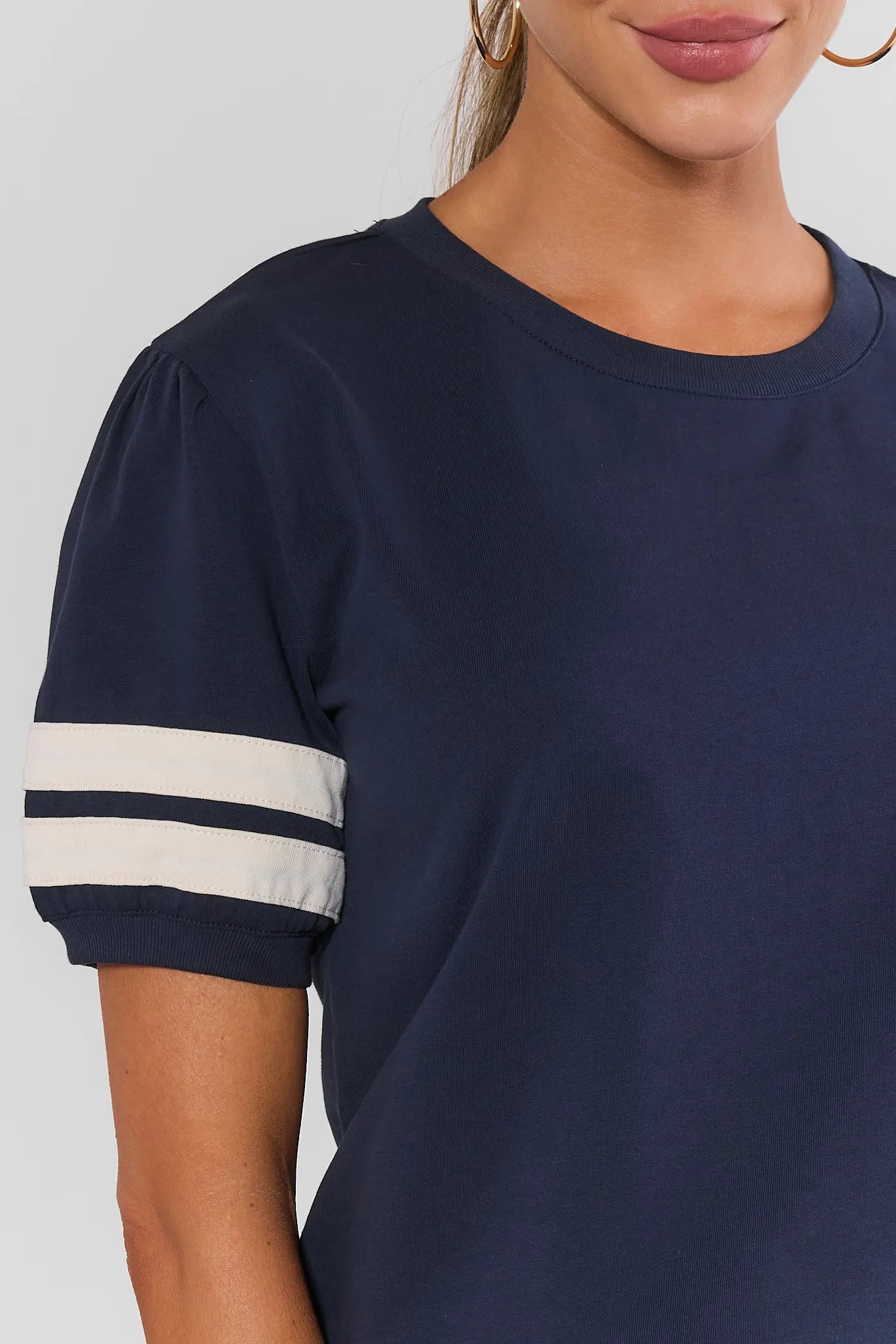 close up A woman modeling a navy blue short-sleeve t-shirt with white stripes on the sleeves, paired with white wide-leg pants and white sneakers, against a gray studio background.