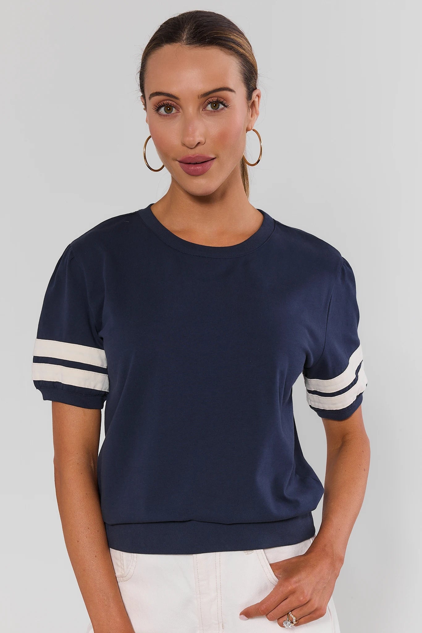 A woman modeling a navy blue short-sleeve t-shirt with white stripes on the sleeves, paired with white pants, against a light gray background.