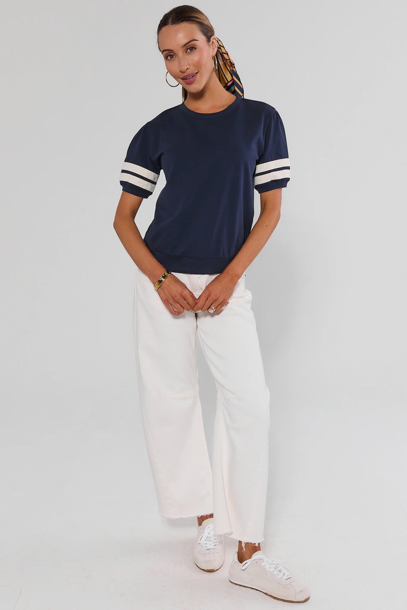 A woman modeling a navy blue short-sleeve t-shirt with white stripes on the sleeves, paired with white wide-leg pants and white sneakers, against a light gray studio background.