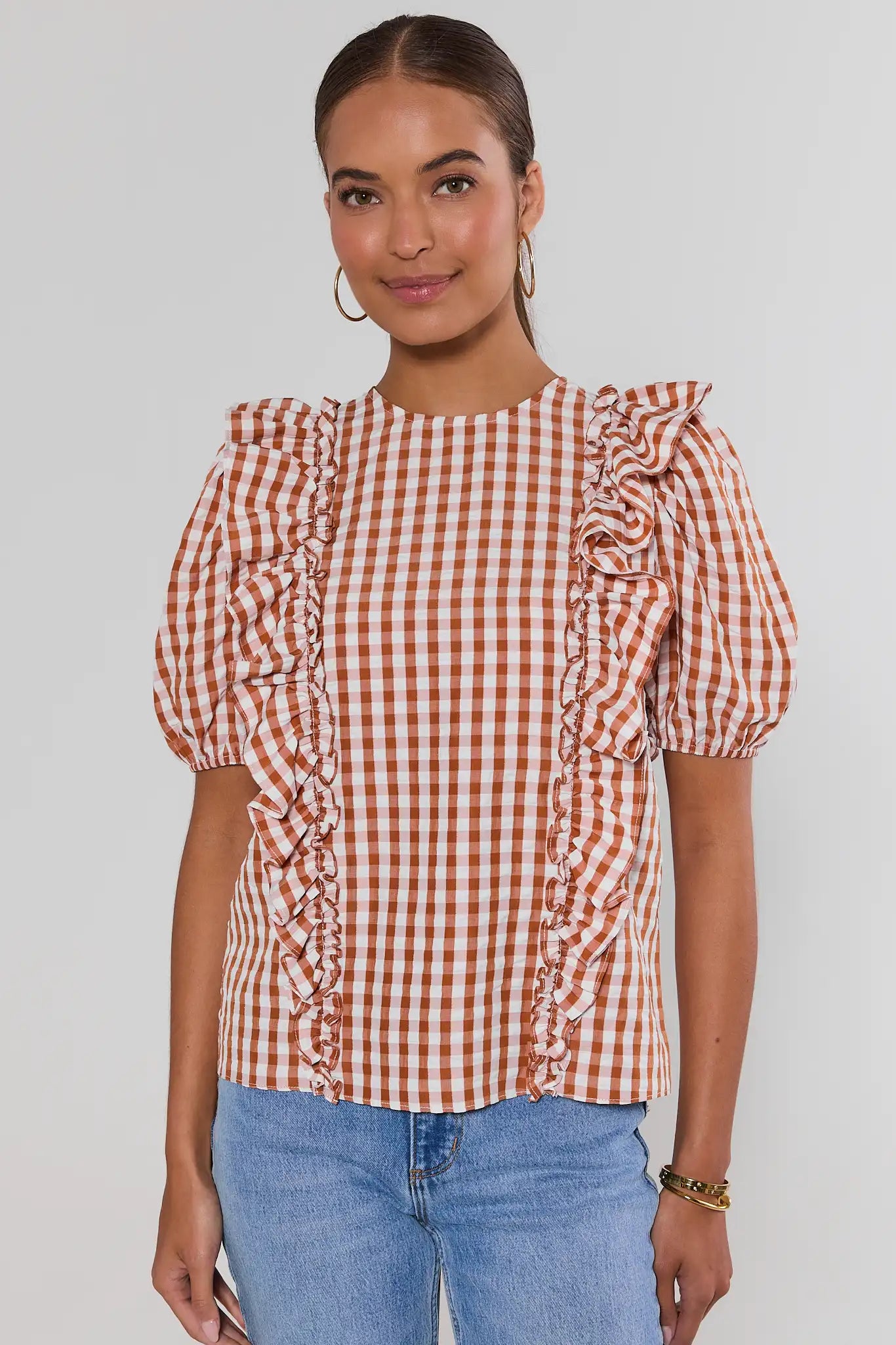 A woman wearing a rust-orange and white gingham check blouse with ruffled details on the shoulders and sides, paired with light blue jeans, against a light studio background.