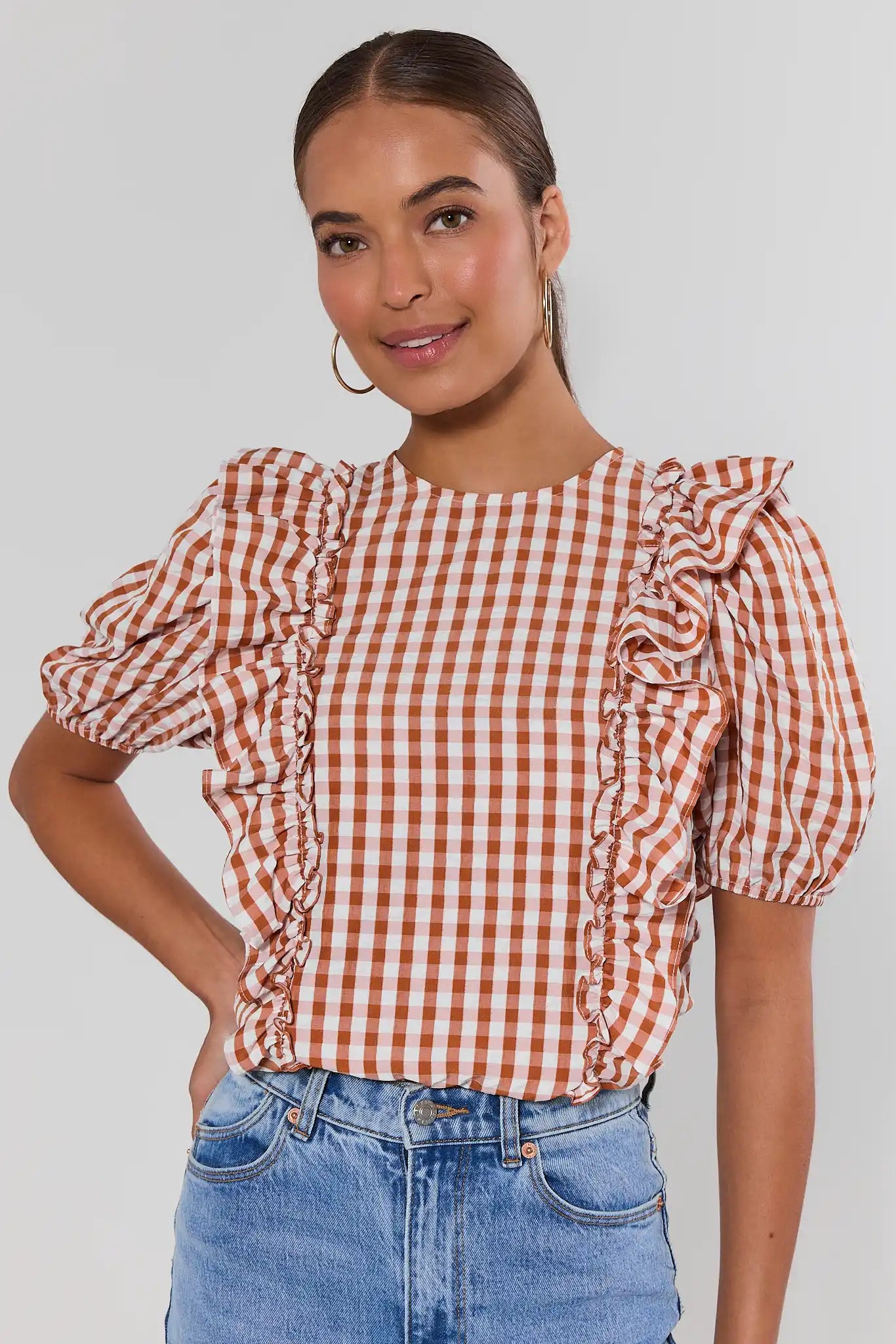 A woman wearing a rust-orange and white gingham short-sleeve blouse with ruffled trim on the sleeves, paired with blue jeans, against a light gray studio background.