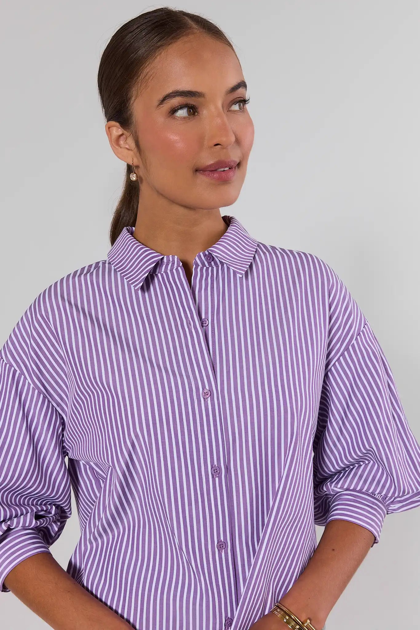 A woman wearing a lavender and white vertically striped button-down shirt with three-quarter sleeves, shown from the chest up against a plain light gray studio background.