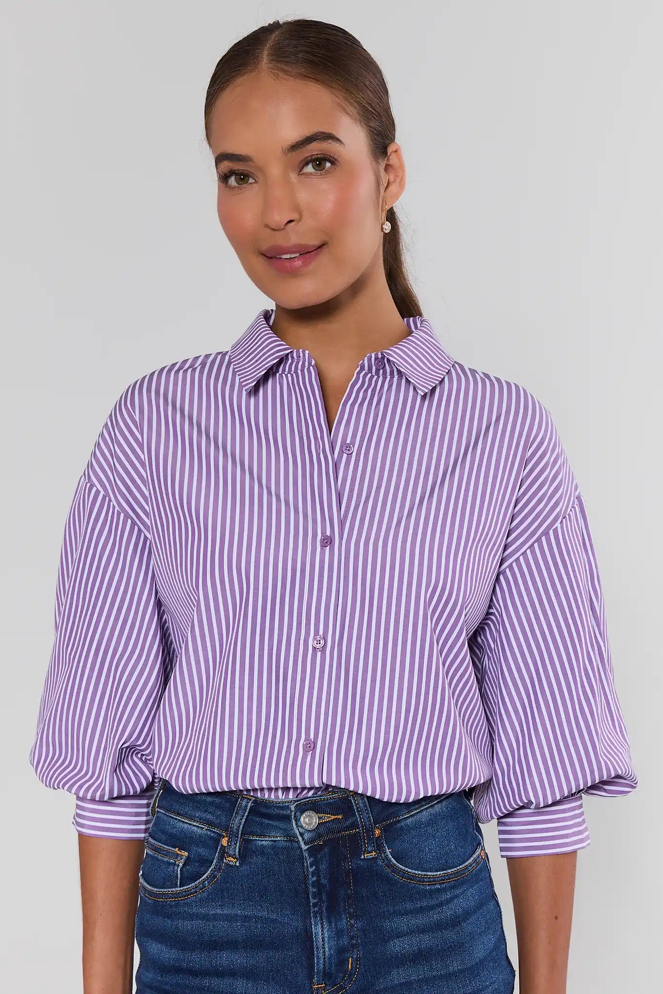 A woman wearing a lavender and white vertically striped button-down shirt with three-quarter sleeves, against a plain light gray studio background.