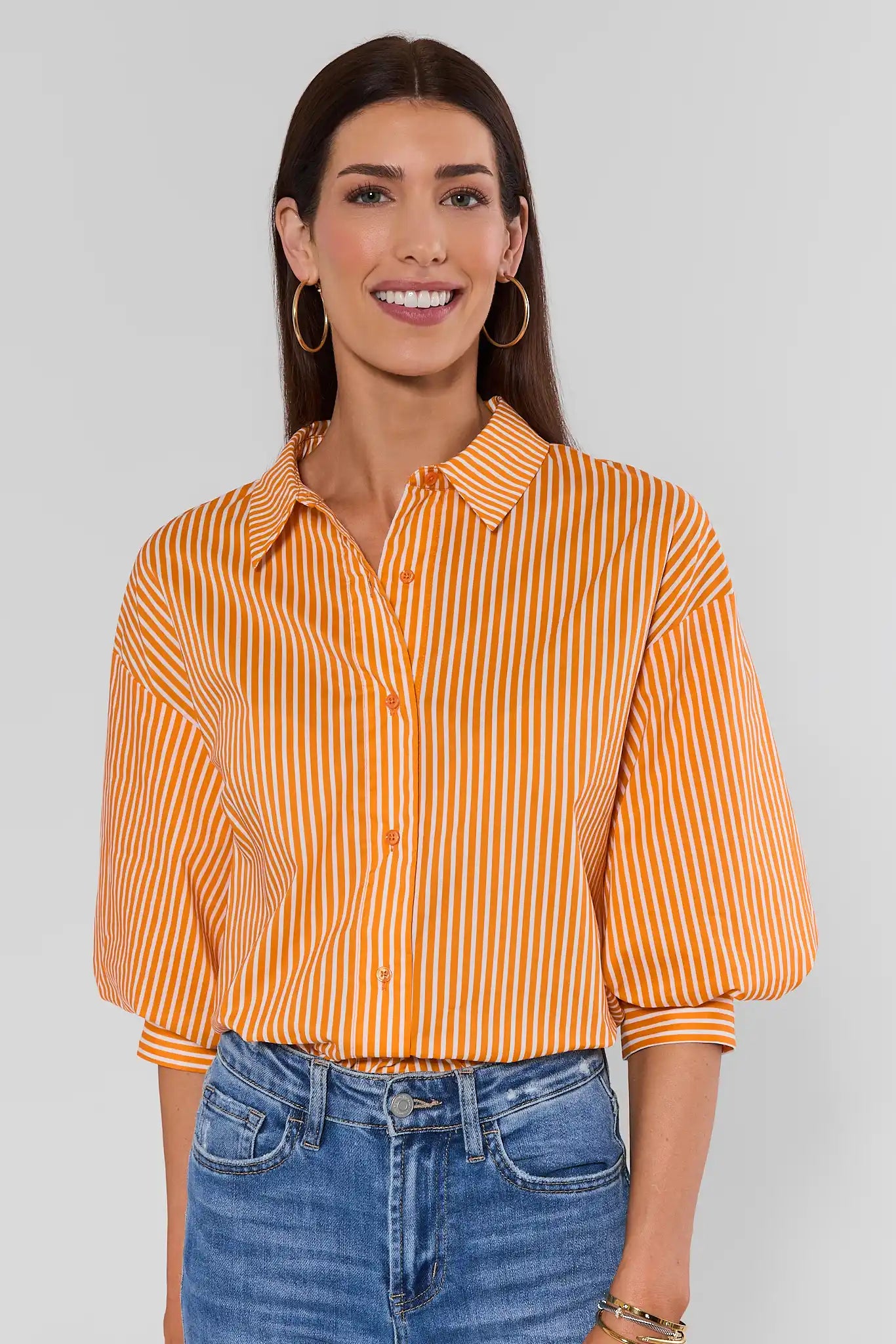 A woman wearing a cropped orange and white vertically striped button-up blouse with puff sleeves, paired with blue jeans, against a solid light background.