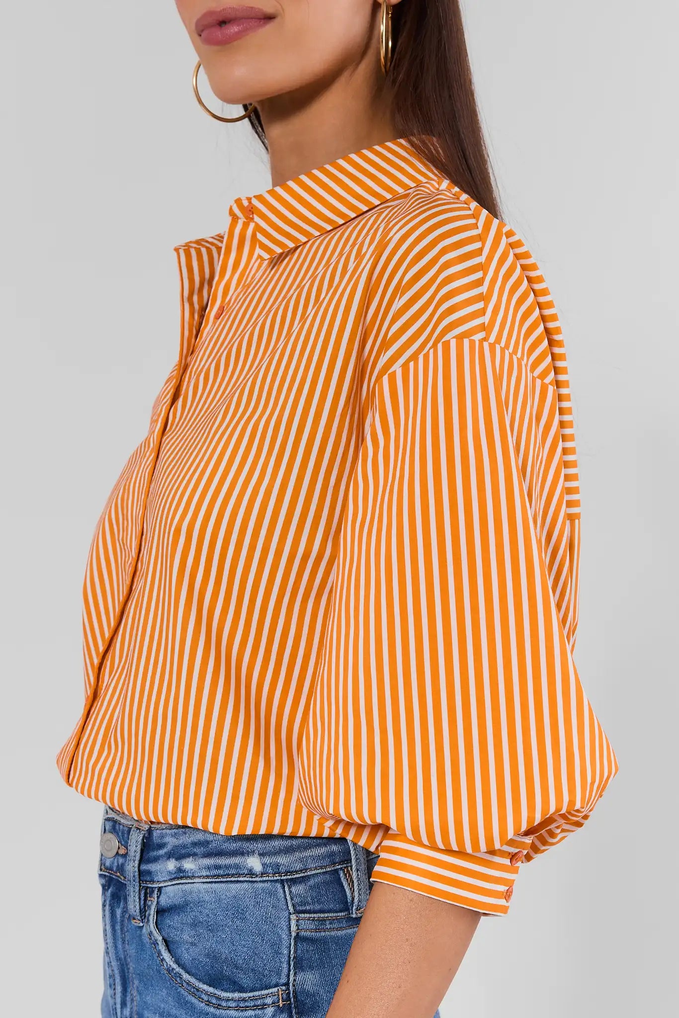 A woman wearing a bright orange and white vertically striped button-up blouse with voluminous elbow-length puff sleeves, paired with blue jeans against a gray studio background.