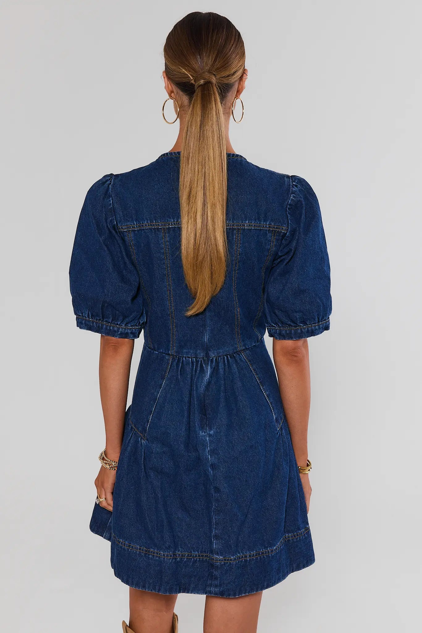 A woman viewed from the back wearing a dark blue denim dress with puff sleeves and a gathered waist, against a plain light gray studio background.