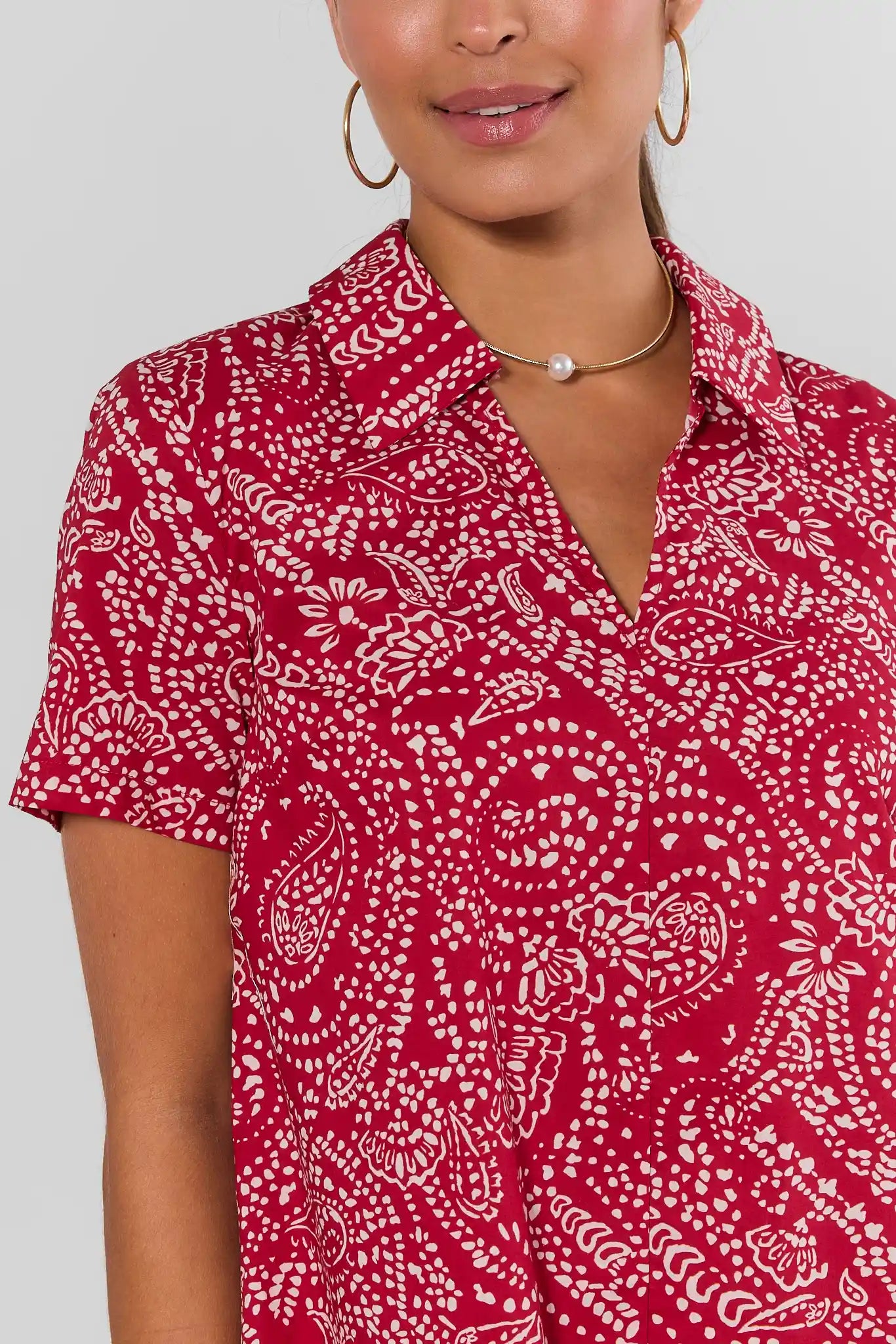 Close up a woman modeling a short-sleeve collared mini dress with a red and white paisley pattern, set against a plain light gray studio background.