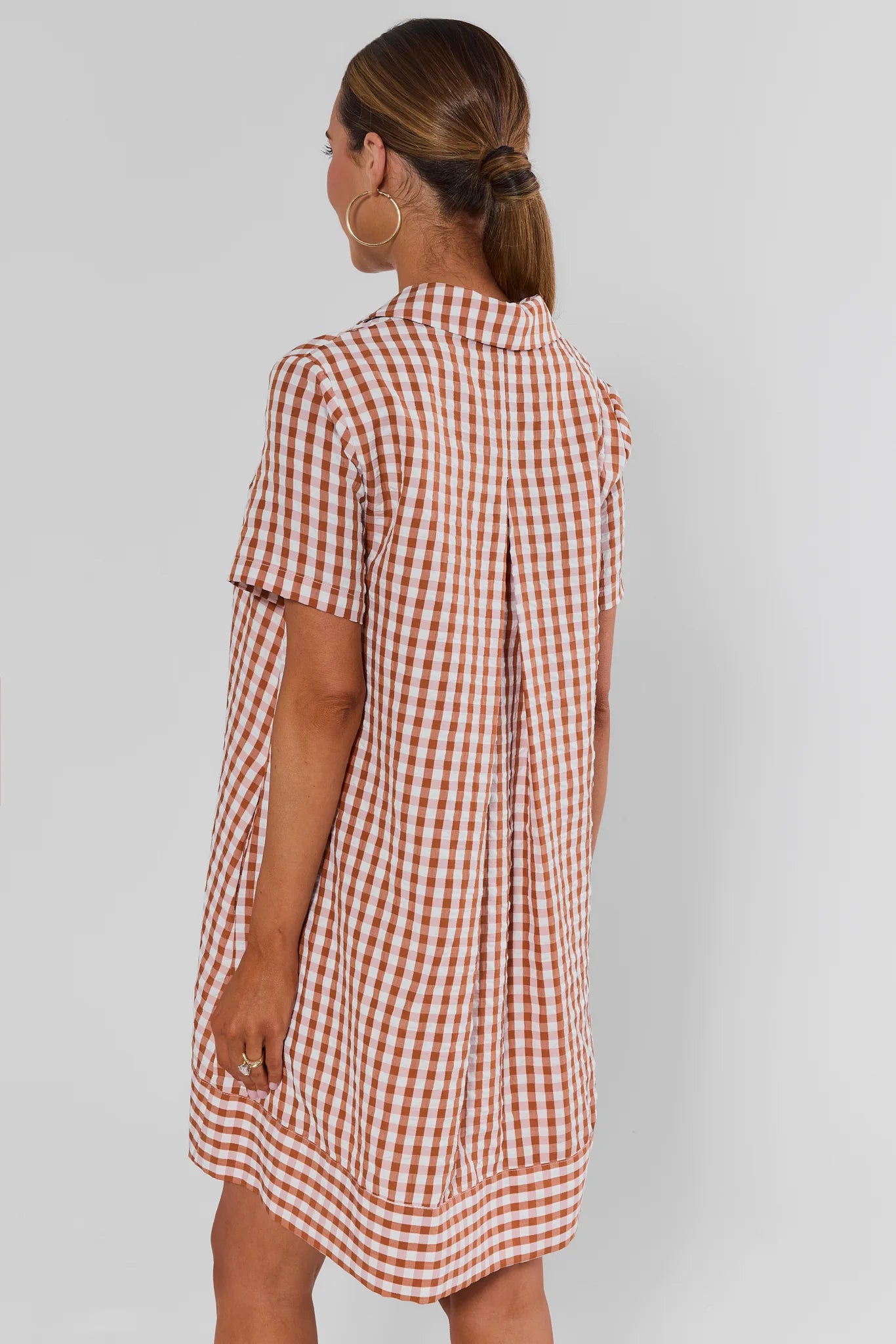 A woman shown from the back wearing a short-sleeve gingham shirt dress in a red and white check pattern, set against a plain light gray studio background.