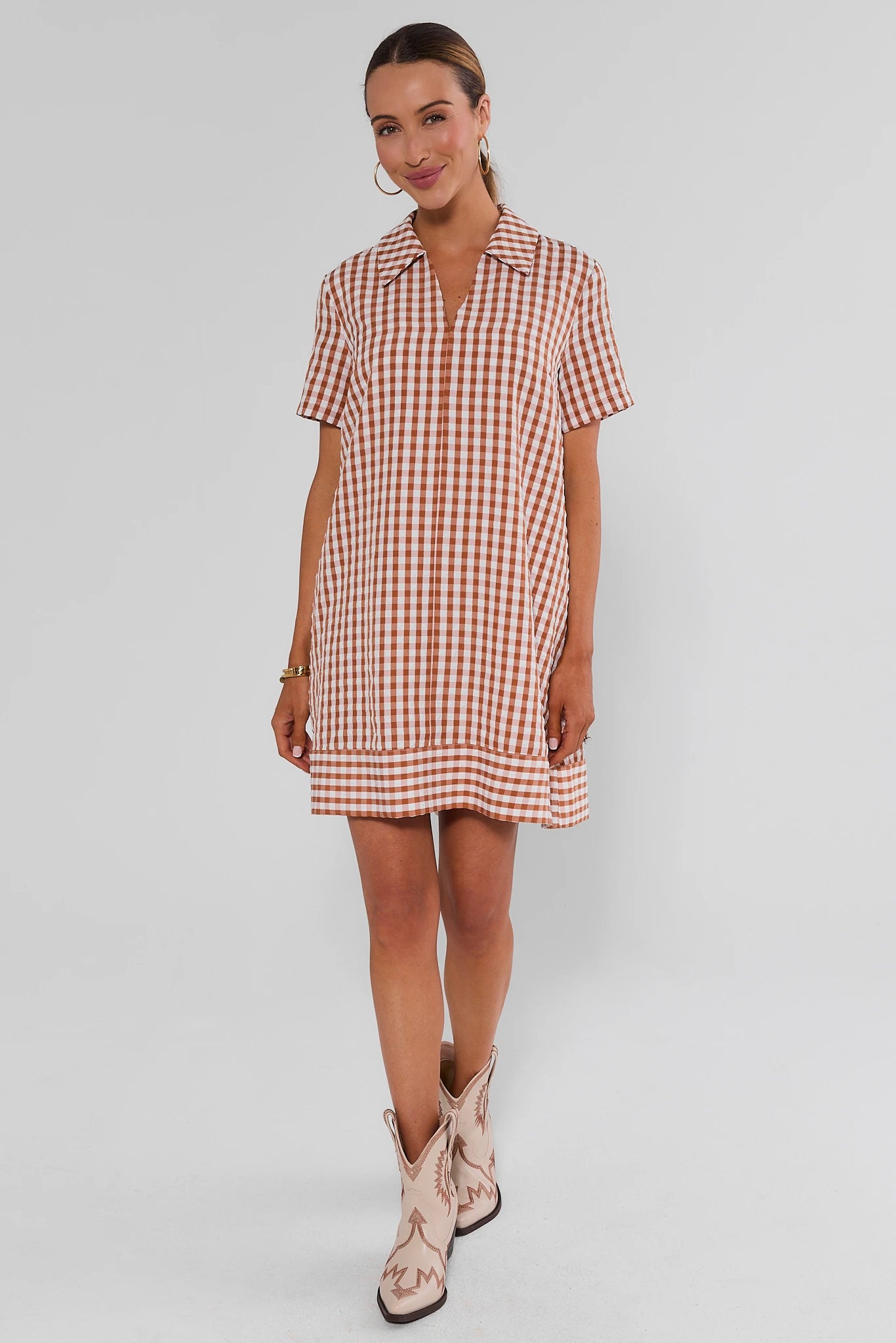 A woman modeling a short-sleeve collared mini dress with a red and white gingham check pattern, paired with light pink cowboy boots, against a light gray studio background.