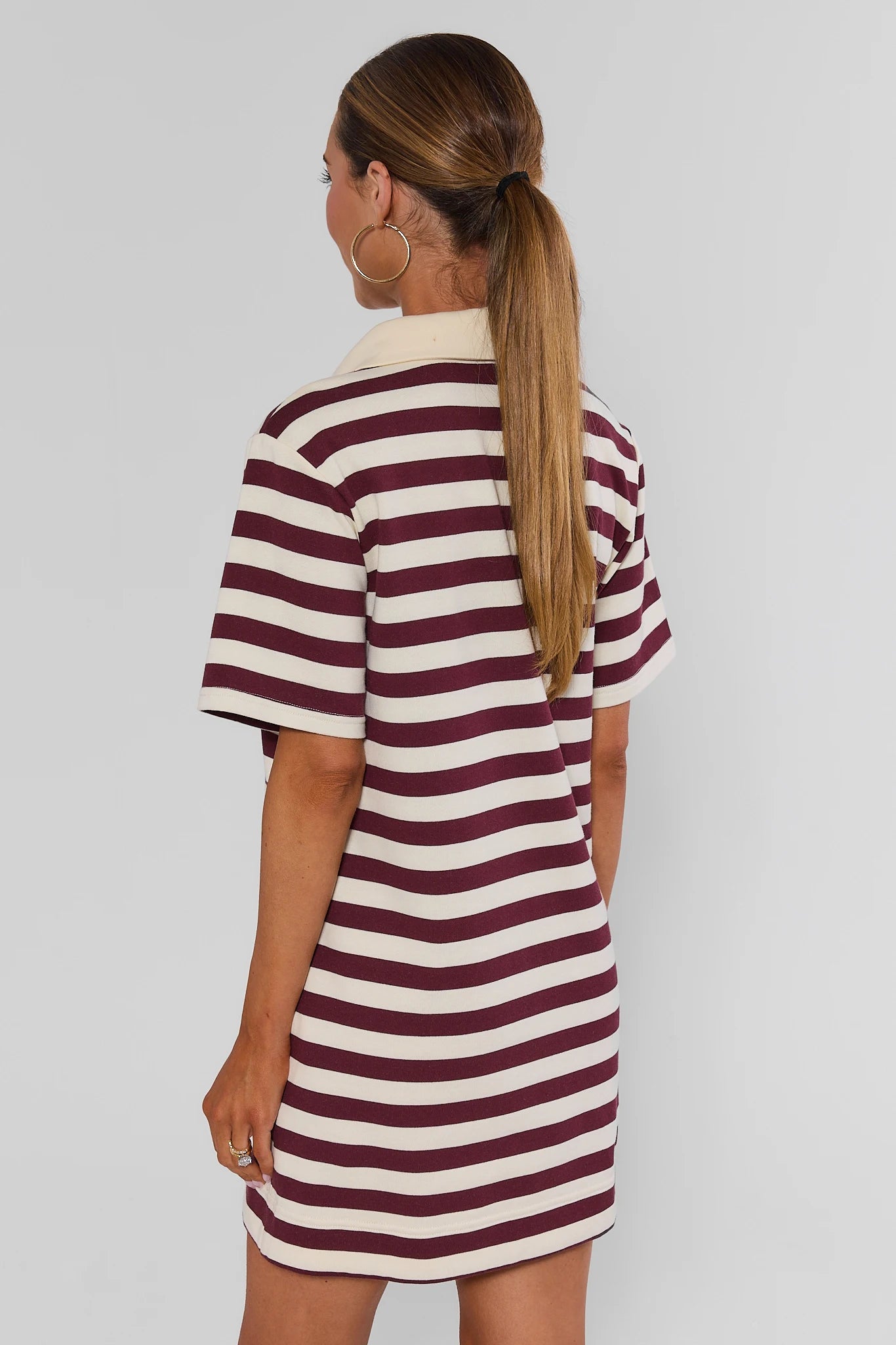 A woman shown from the back wearing a short-sleeve polo dress with horizontal burgundy and cream stripes, set against a plain light gray studio background.