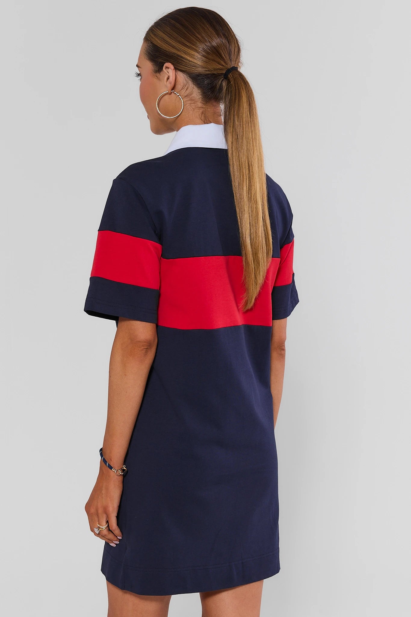 A woman shown from the back wearing a short-sleeve colorblock polo dress with navy blue, red, and white horizontal stripes, set against a plain light gray background.
