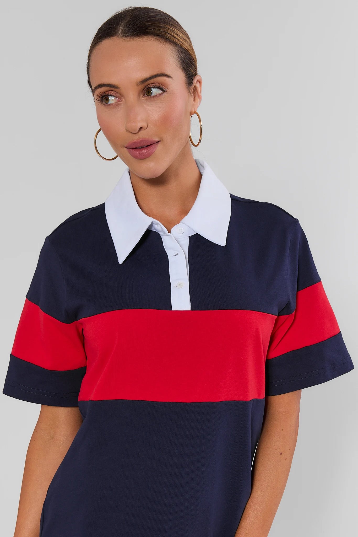 Close up a woman wearing a short-sleeve colorblock polo shirt with navy blue, red, and white stripes, paired with gold hoop earrings, against a plain light gray background.