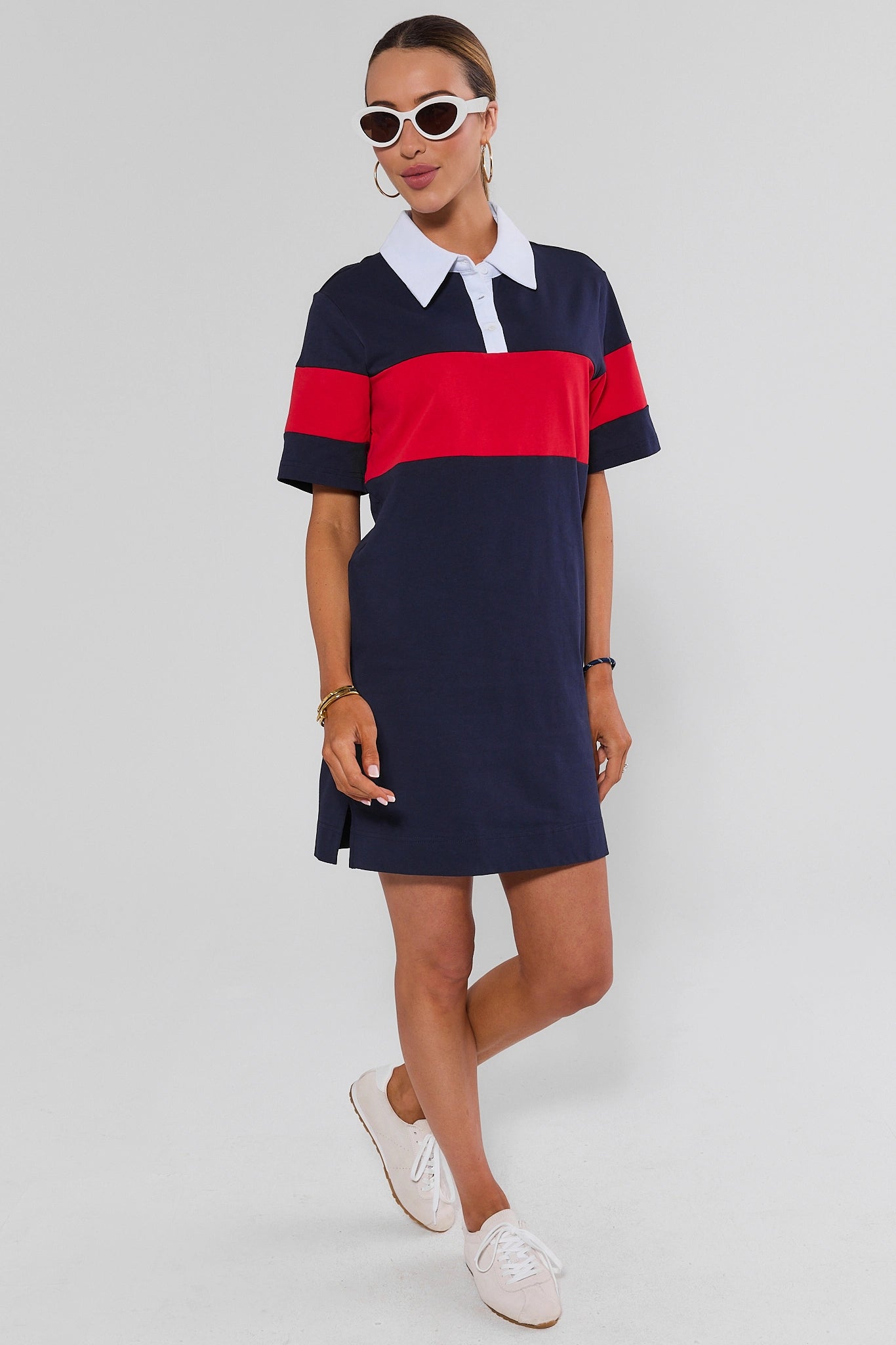 A woman modeling a short-sleeve polo dress with a navy blue base, a bold red horizontal stripe across the chest and sleeves, and a white collar, paired with white sneakers against a light gray studio background.
