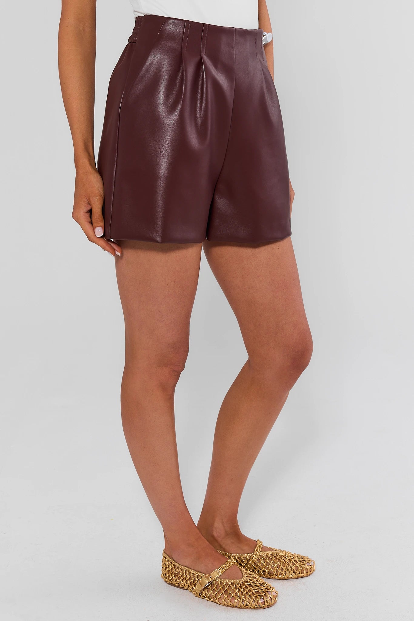 A model wearing high-waisted, pleated faux-leather shorts in a deep burgundy color, paired with woven flat shoes, against a light background.