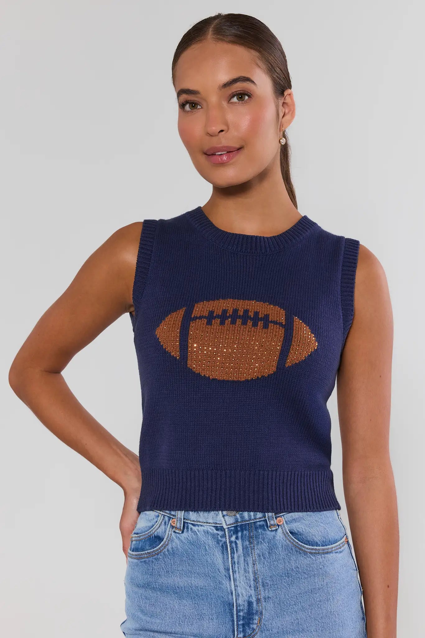 A woman wearing a navy blue sleeveless knit sweater with a brown football graphic on the front, paired with light blue denim jeans, against a light gray studio background.
