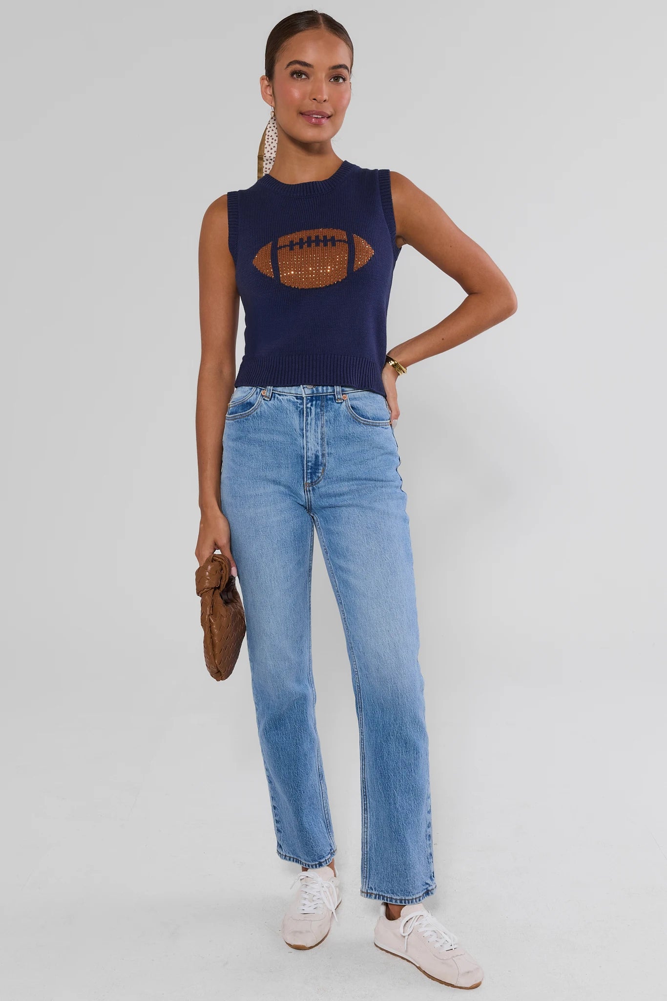A woman wearing a navy blue sleeveless knit sweater with a large brown football graphic on the front, with light wash jeans, shown against a light gray studio background