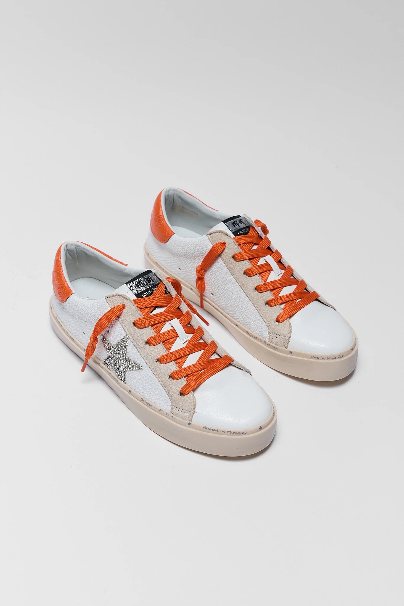 A pair of white leather low-top sneakers with vibrant orange laces, orange heel tab accents, and a distressed silver star logo on the side, displayed on a light gray background.