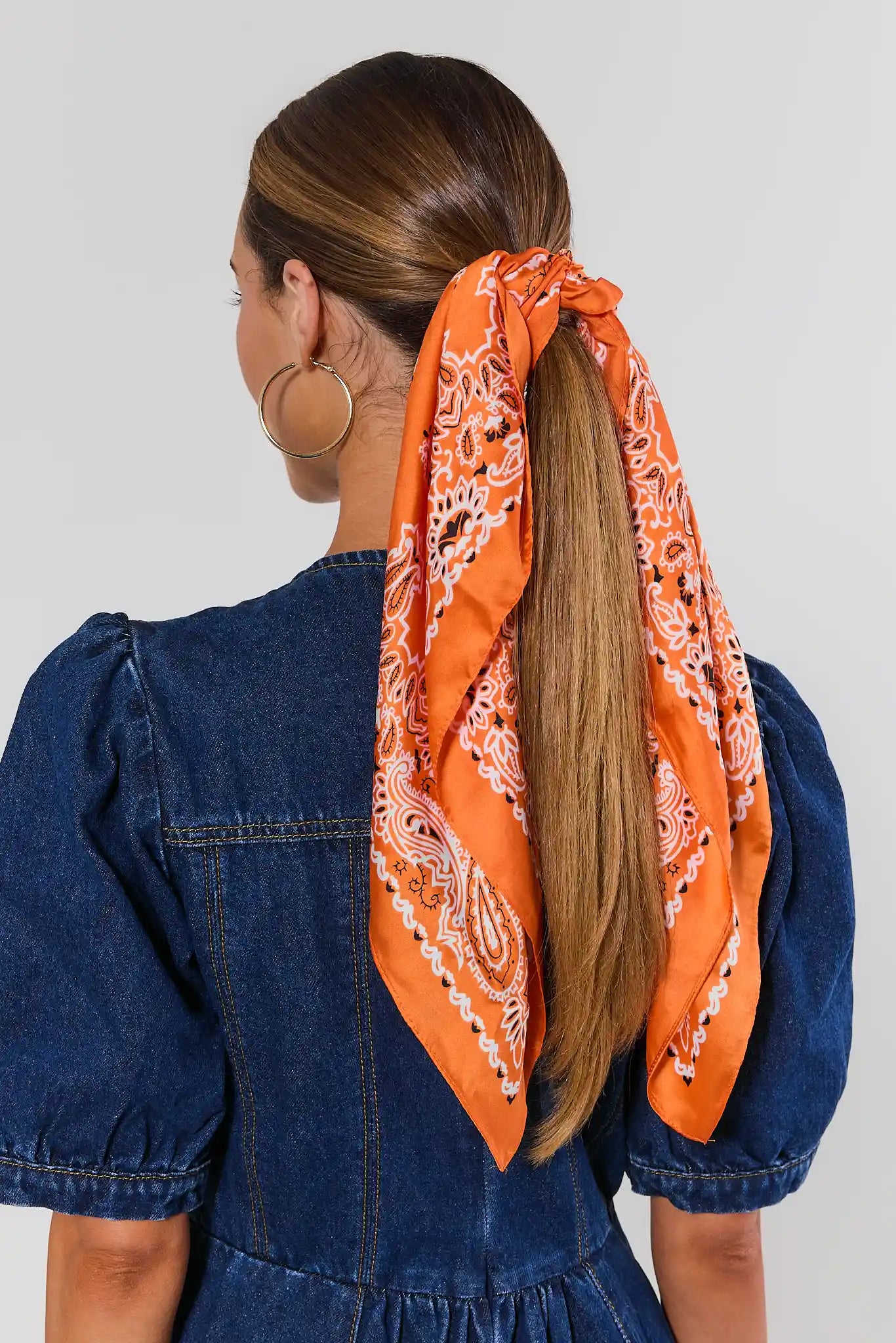 An orange paisley bandana tied around a woman's ponytail, shown from the back. The model is wearing a blue denim puff-sleeve dress against a light gray studio background.