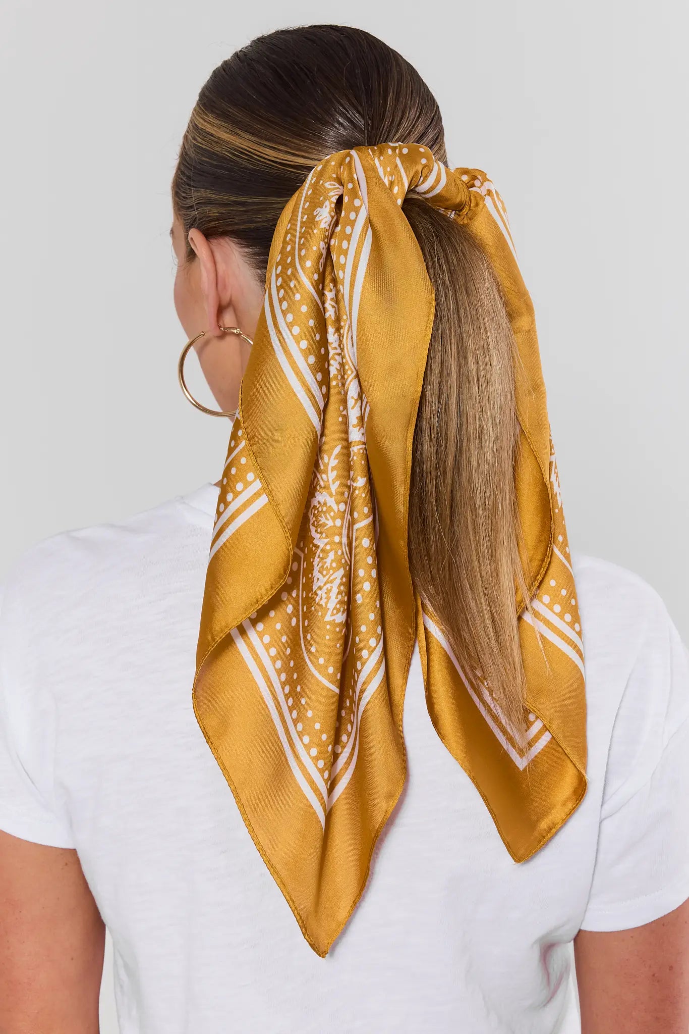 A woman shown from the back wearing a white t-shirt and large hoop earrings, with her hair tied up in a ponytail using a mustard yellow satin bandana with a white paisley pattern.