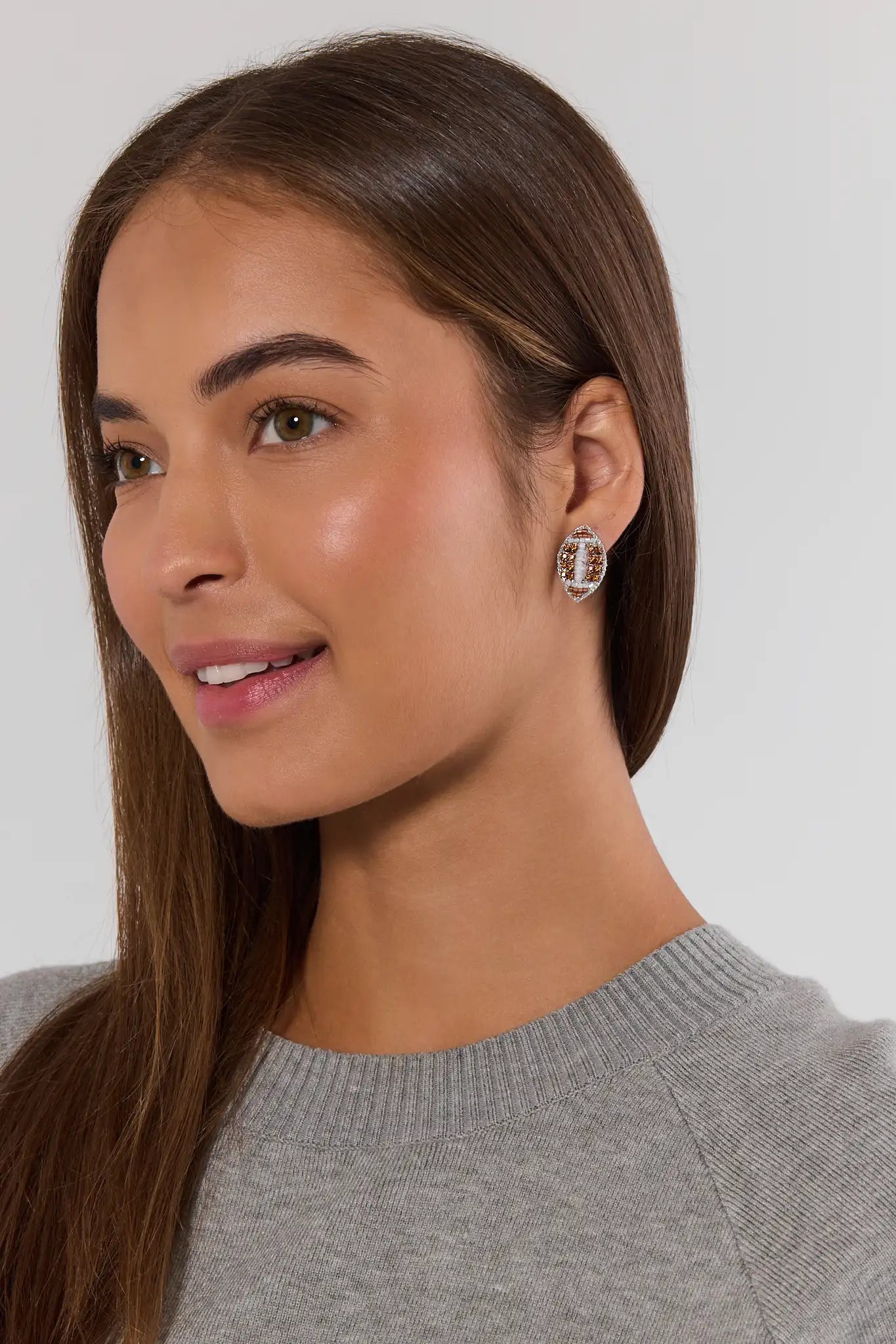 A close-up of a woman with long brown hair wearing a gray knit sweater and a small silver hoop earring with clear stones, set against a plain light gray background.