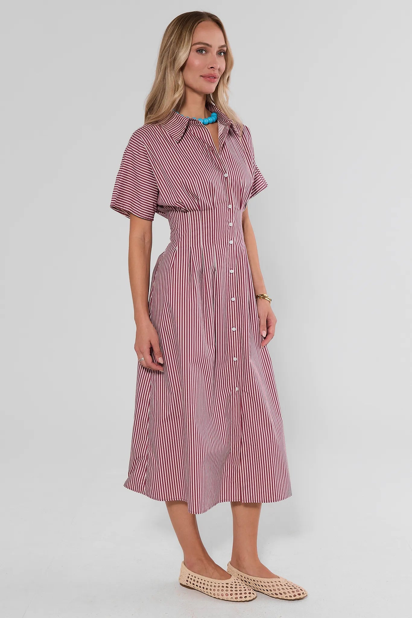A woman modeling a short-sleeve midi shirt dress with vertical pink and white stripes, a button-down front, and a pointed collar, photographed against a light gray studio background.