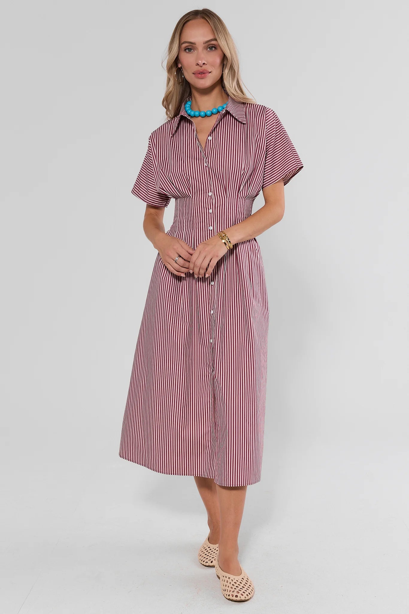 A woman modeling a short-sleeve midi shirt dress with vertical pink and white stripes, featuring a button-down front and collar, set against a plain light gray studio background.