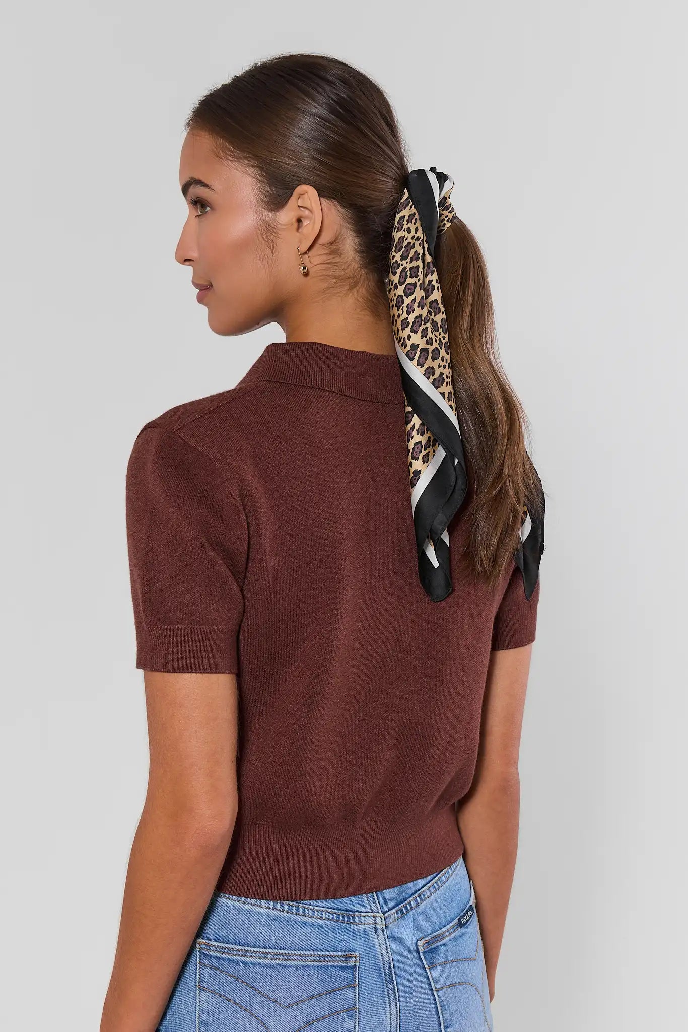 A woman shown from the back wearing a short-sleeve ribbed knit polo shirt in a deep burgundy brown, paired with light blue jeans. Her hair is tied back with a leopard-print and black scarf.