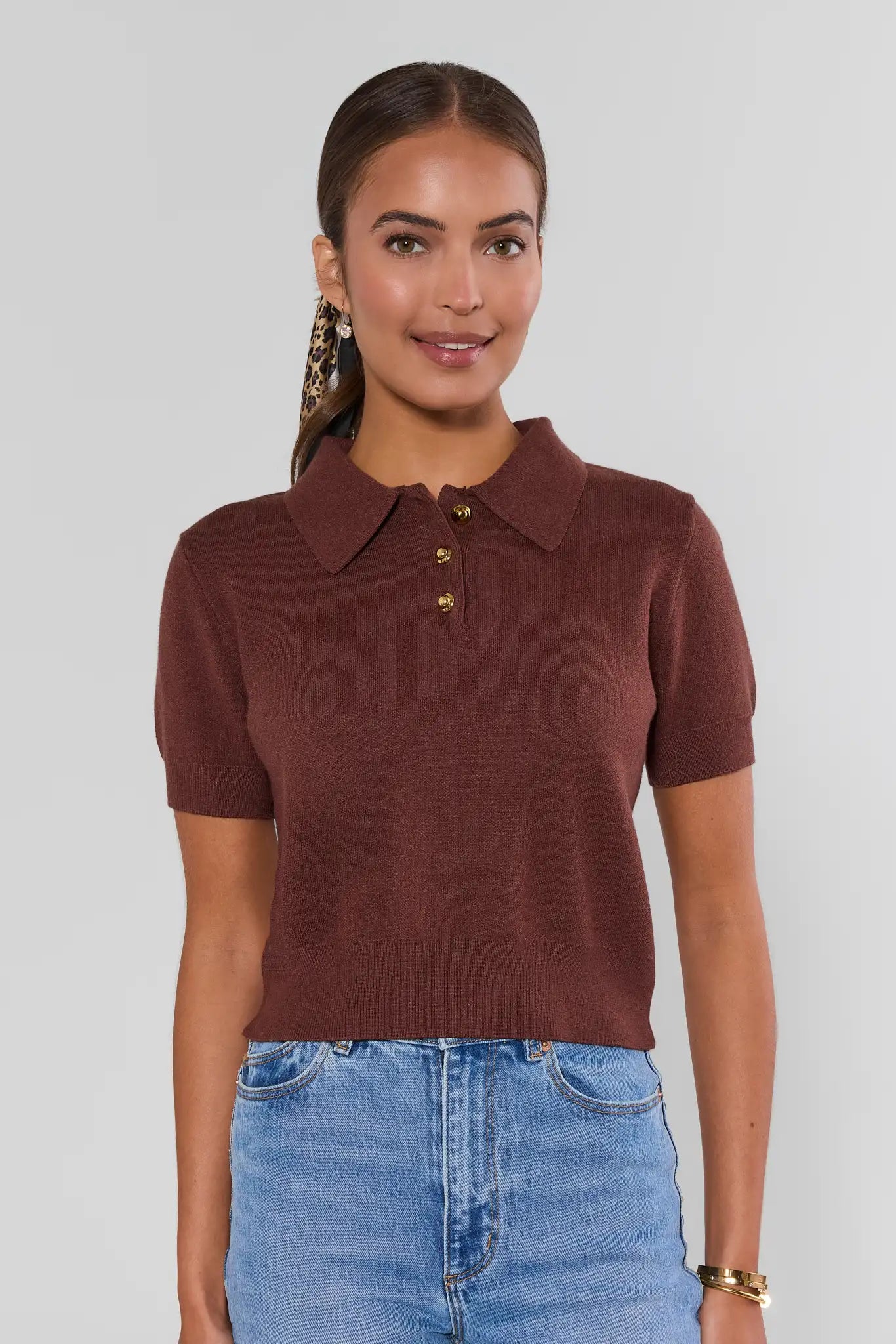 A woman wearing a short-sleeve knit polo shirt in a deep rust brown color, paired with light blue jeans, against a solid light gray background.